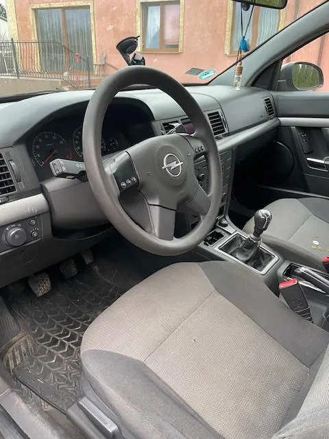 Wnętrze samochodu z widocznym kierownicą z logo Opel, deską rozdzielczą z wskaźnikami prędkości i obrotomierzem, dźwignią zmiany biegów oraz szarymi siedzeniami. Na podłodze znajdują się czarne dywaniki. Przez przednią szybę widoczny jest ceglany budynek z oknami i balkonem.