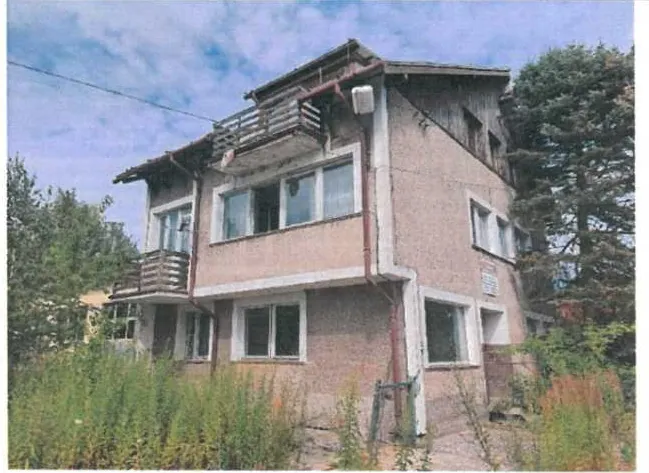 Dwupiętrowy dom o różowawym kolorze elewacji z białymi obramowaniami okien. Na pierwszym piętrze znajduje się balkon z drewnianym ogrodzeniem, a na dachu widoczne są elementy konstrukcyjne i rynny. Dom otoczony jest wysoką trawą i drzewami. Niebo na tle obrazu jest częściowo zachmurzone. Brak czytelnego tekstu.