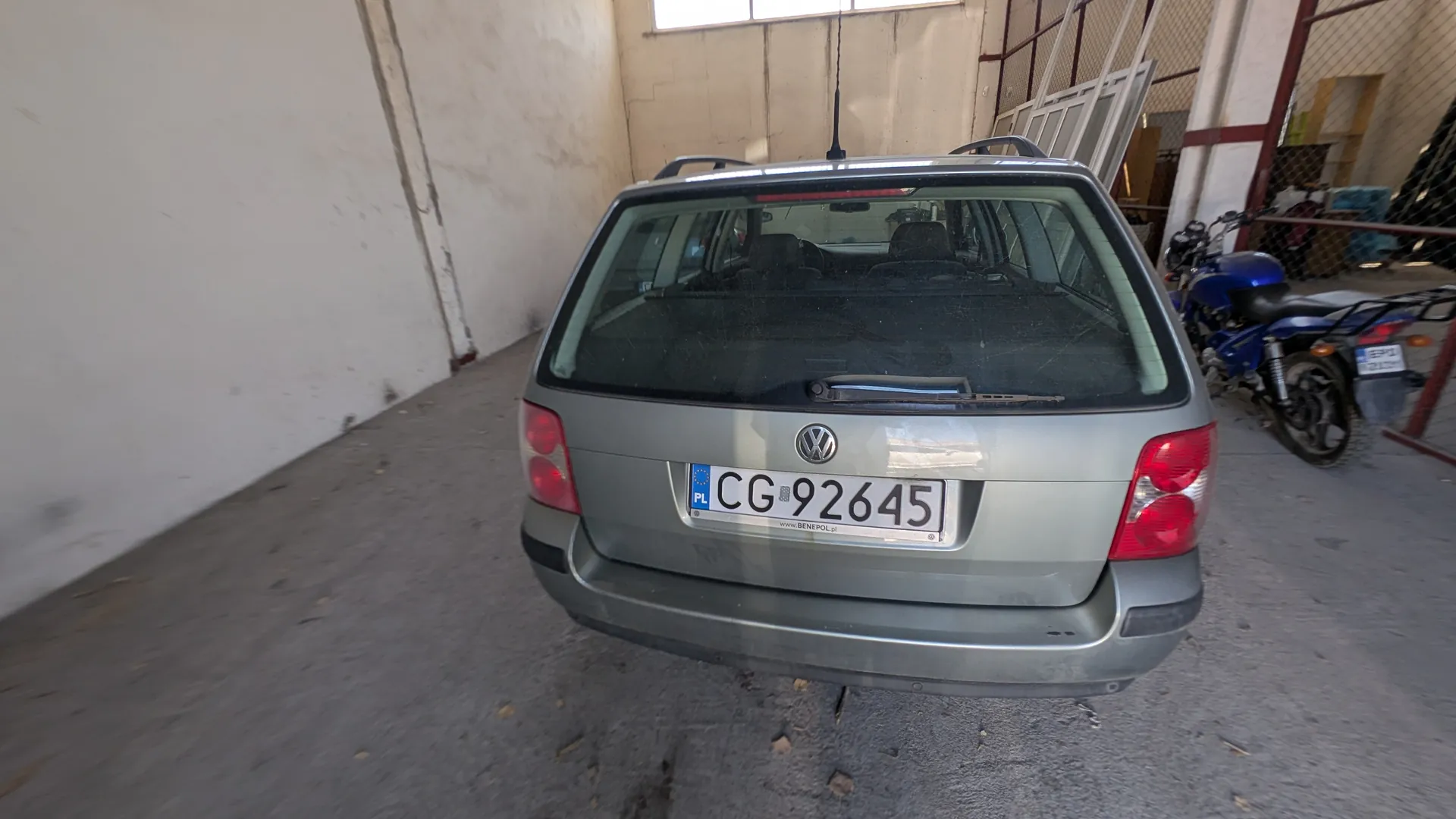 Z tyłu widoczny szary samochód osobowy marki Volkswagen z tablicą rejestracyjną CG 92645 oraz oznaczeniem PL. Po prawej stronie stoi niebieski motocykl. Scena rozgrywa się w pomieszczeniu przemysłowym z betonowymi ścianami, oknem w tle i betonową podłogą z rozrzuconymi liśćmi.