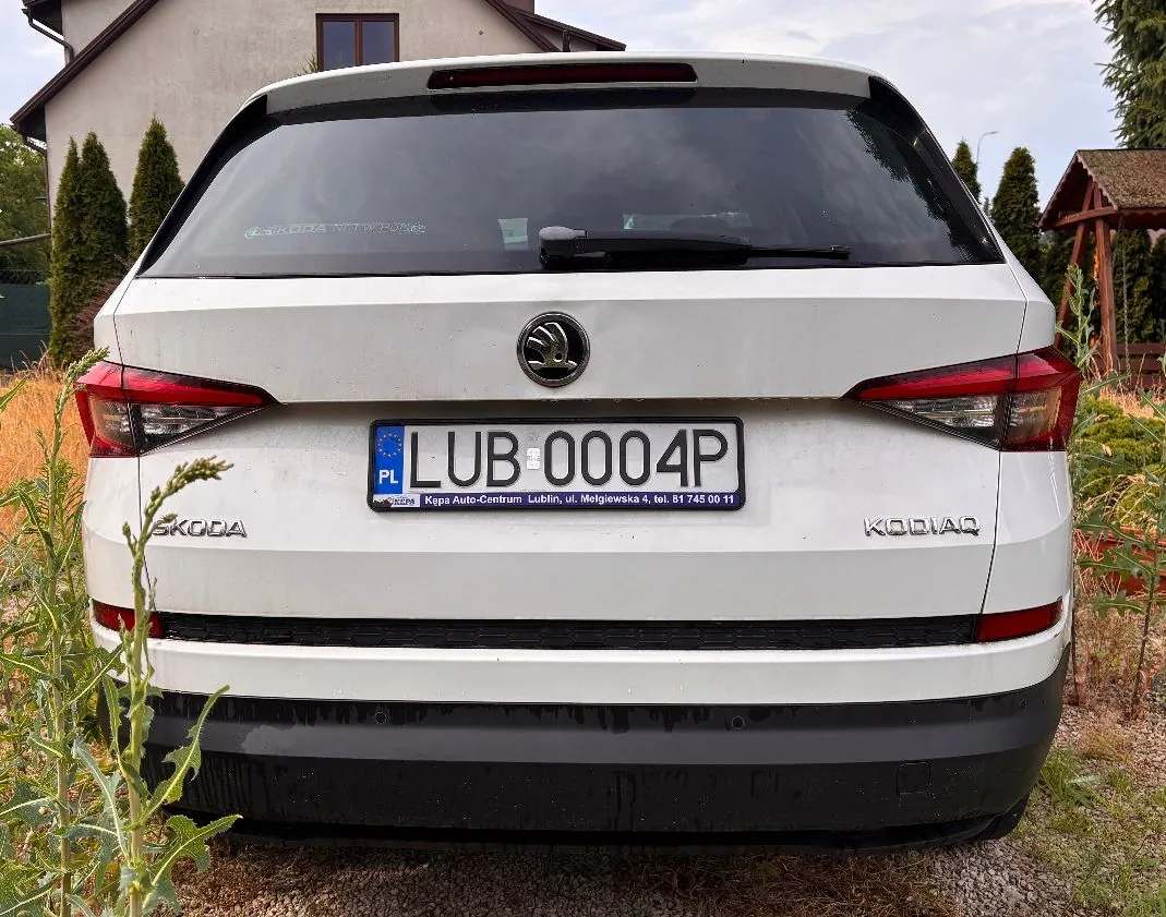 Tył białego samochodu marki Škoda zaparkowanego przed domem. Na samochodzie widoczna jest tablica rejestracyjna z napisem 'LU B004P'. W tle widać fragmenty budynków oraz zielone krzewy i drzewa.