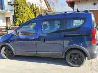 Niebieski samochód typu minivan zaparkowany na jasnym podjeździe. Pojazd jest pokazany z profilu od strony pasażera. W tle widoczne są zielone krzewy oraz fragmenty budynków mieszkalnych.