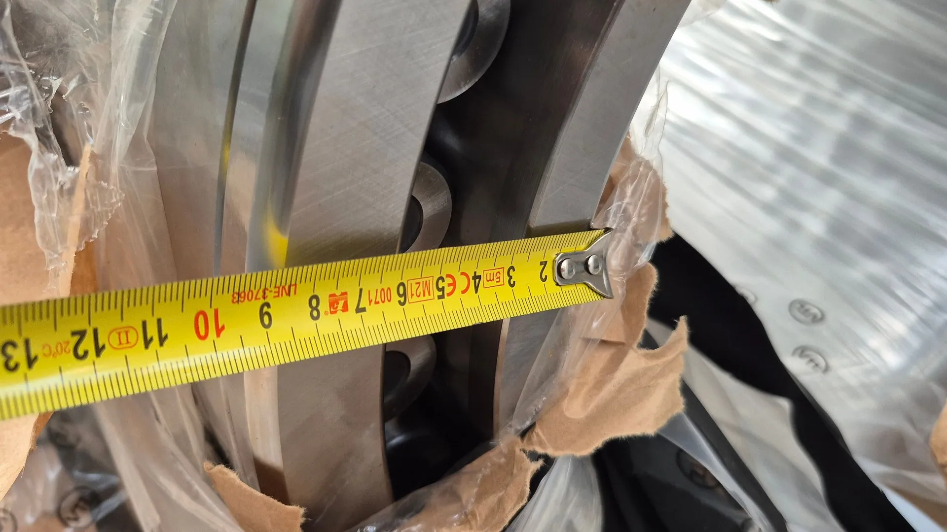 Zbliżenie na metalowy element mechaniczny, prawdopodobnie łożysko lub koło zamachowe, częściowo owinięte w przezroczystą folię i brązowy papier ochronny. Przez środek obrazu przebiega poziomo żółta miara zwijana, wskazująca wymiar elementu. Na mierze widoczne są cyfry od 9 do 13 oraz oznaczenia techniczne: '3 5m 4CE5 M21 6 0071 7 LNE 37053'.