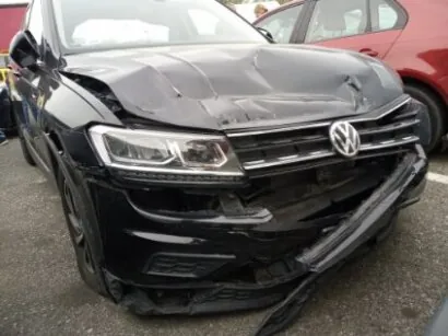 Zdjęcie przedstawia czarny samochód marki Volkswagen z widocznymi uszkodzeniami na przodzie. Maskownica i zderzak są mocno zdeformowane, a część zderzaka zwisa. W tle widoczny jest czerwony samochód. Scena rozgrywa się na otwartym placu parkingowym lub ulicy.