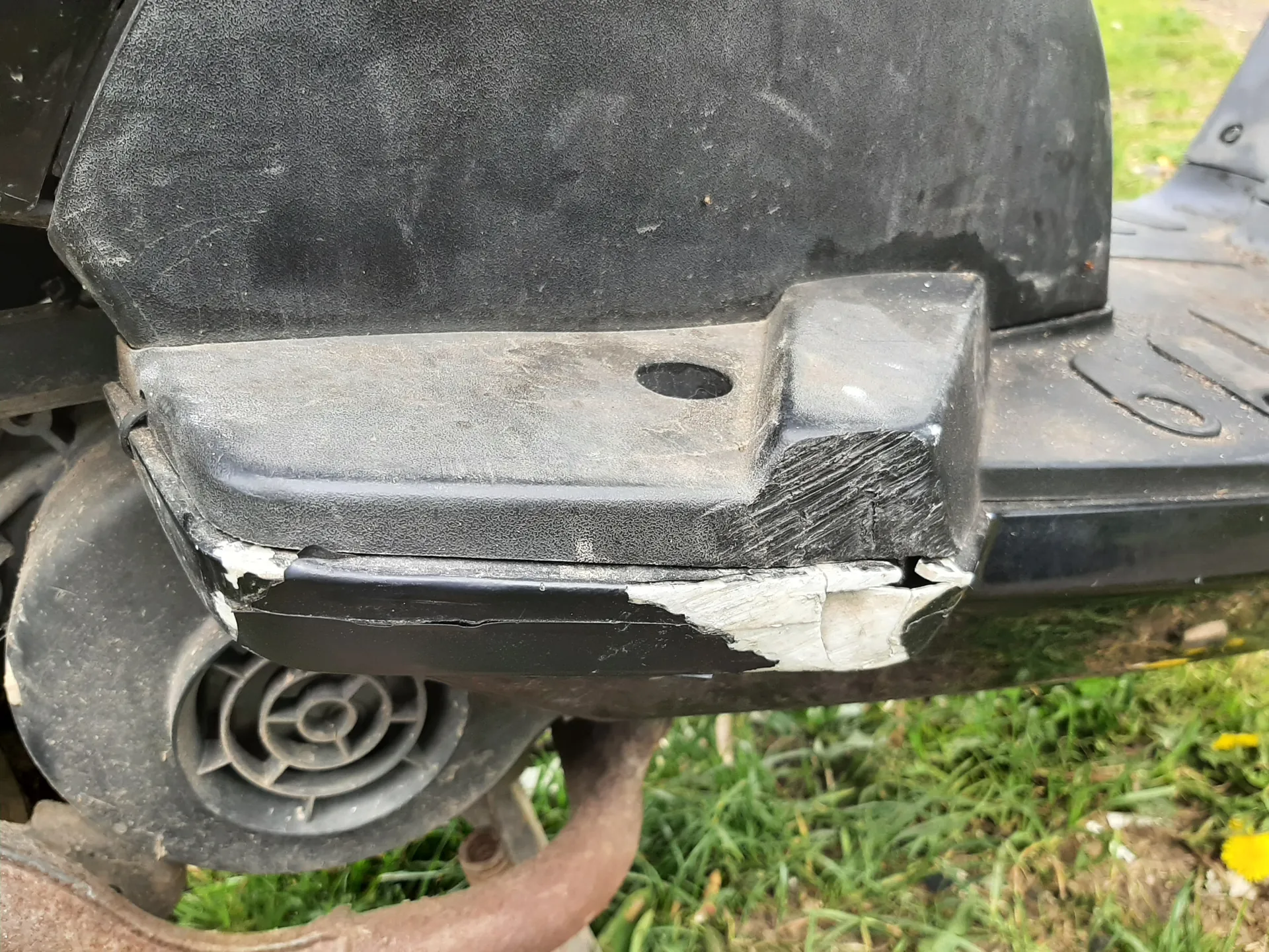 Zbliżenie na uszkodzoną czarną plastikową część pojazdu lub maszyny z widocznymi zadrapaniami, pęknięciami i odłupanym białym materiałem. W tle zielona trawa z żółtymi kwiatami oraz okrągły element wentylacyjny. Brak czytelnego tekstu.