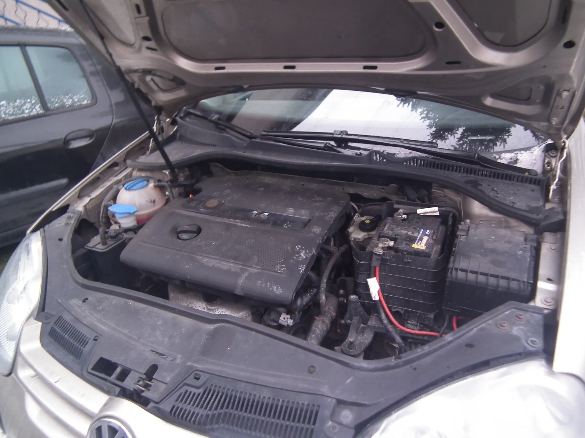 Samochód marki Volkswagen z otwartą maską, ukazujący komorę silnika. W komorze widoczne są czarne elementy silnika, dwa niebieskie korki (prawdopodobnie zbiornik chłodniczy i płukacz szyby), akumulator z czerwonymi i czarnymi przewodami oraz napis 1.4 na pokrywie silnika. W tle częściowo widoczny czarny samochód i metalowe ogrodzenie. Całość pokryta jest kurzem.