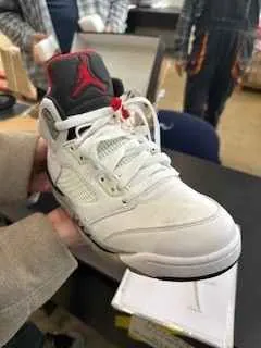 Biała buty sportowe z czerwonym logo na języku i czerwonymi akcentami na sznurówkach, trzymane w dłoni osoby w beżowym swetrze. W tle widoczne są nieostre postacie ludzi oraz białe pudełko na czarnym stole.