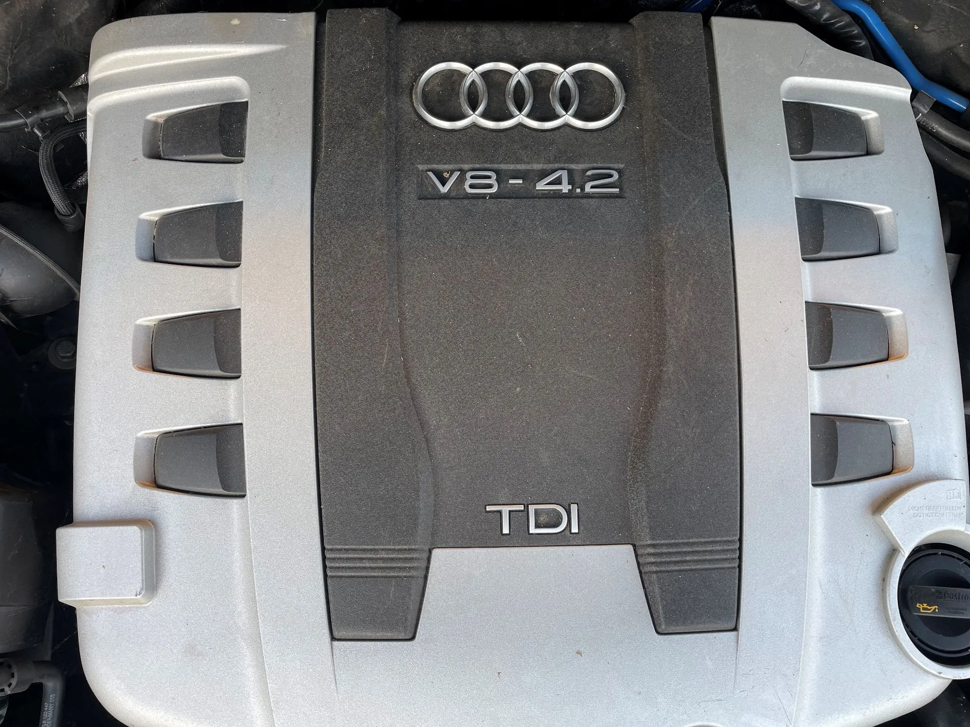 Zbliżenie na górną pokrywę silnika samochodu marki Audi. Pokrywa jest dwukolorowa: srebrna na bokach i czarna w centralnej części. Na górze znajduje się srebrne logo Audi (cztery splecione pierścienie), poniżej napis 'V8-4.2', a w dolnej części czarnego elementu napis 'TDI'. Po prawej stronie widoczny jest żółty korek wlewu oleju.