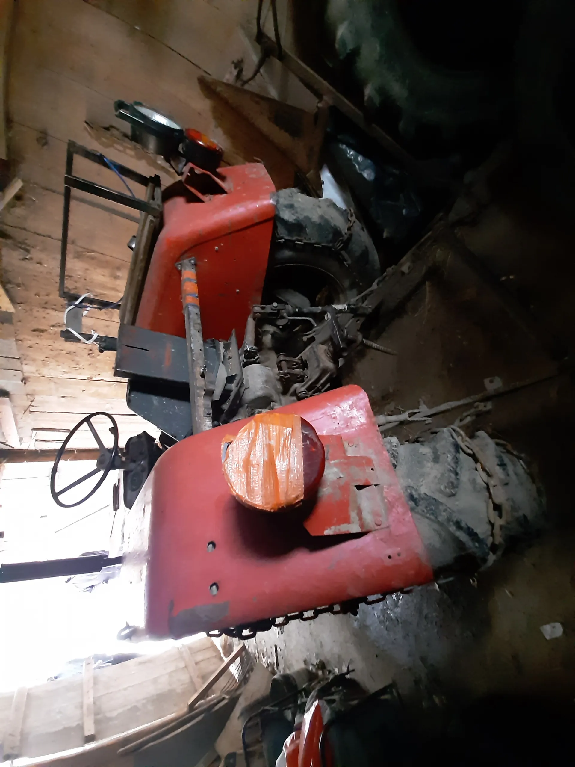 Stary czerwony traktor w drewnianym garażu. Widoczna kierownica, reflektor owinięty pomarańczową taśmą, metalowe części i brudne opony. W tle drewniane ściany, otwarte drzwi wpuszczające światło oraz rozrzucone przedmioty na podłodze.
