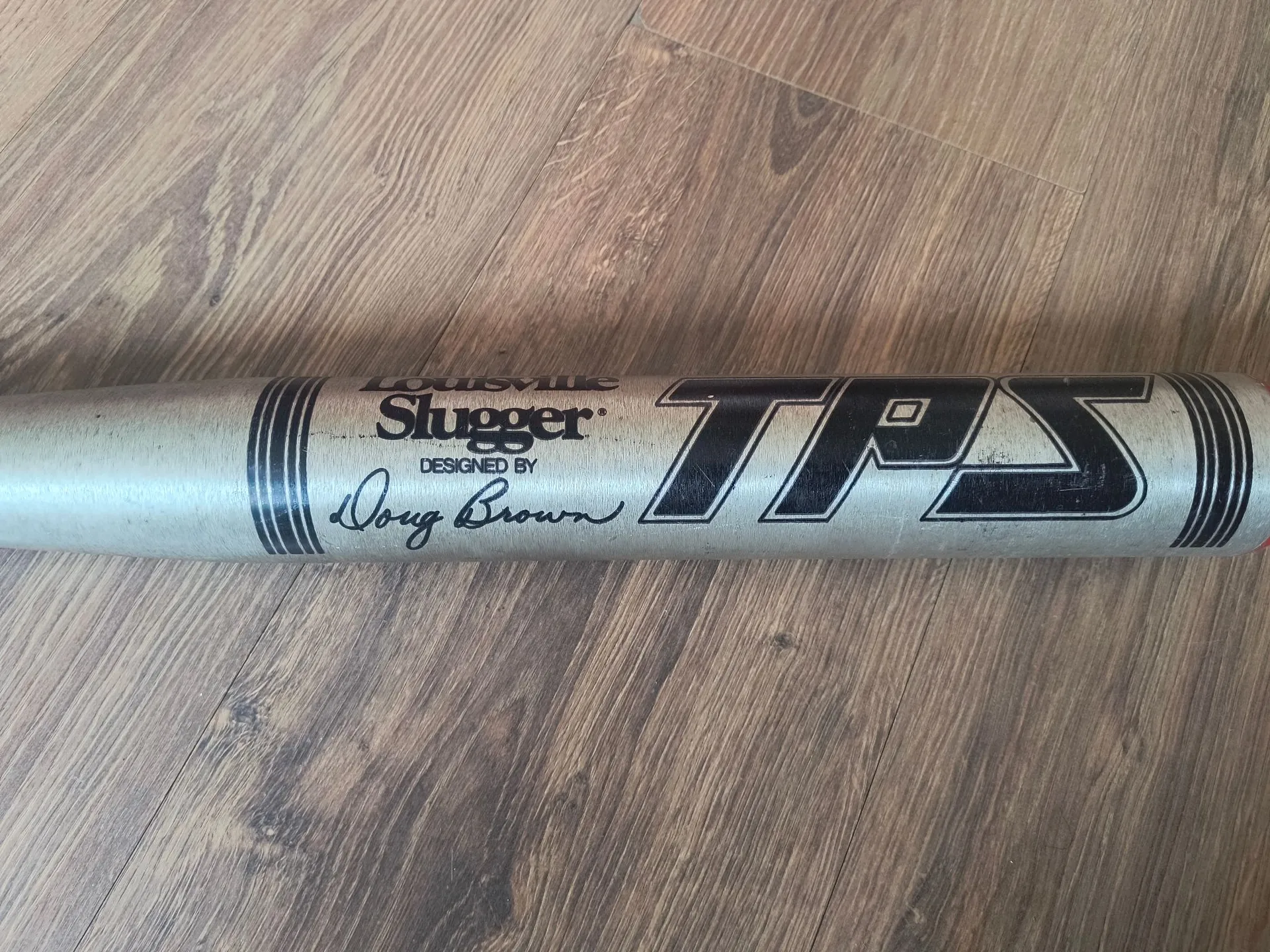 Srebrny kij bejsbolowy leżący poziomo na drewnianej podłodze. Na kiju widnieje czarny napis 'Louisville Slugger', poniżej mniejszy tekst 'DESIGNED BY' oraz podpis 'Doug Brown'. Po prawej stronie znajduje się duży, stylizowany napis 'TPS'.