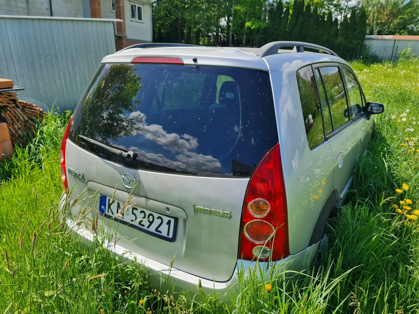Srebrny samochód Mazda Premacy zaparkowany na wysokiej trawie z żółtymi kwiatami. Widok od tyłu z tablicą rejestracyjną PL KM 52921, logo Mazdy i napisem 'mazda' na bagażniku. W tle metalowy płot, stos cegieł, dom z oknami oraz drzewa. W odbiciu na tylnej szybie widoczne chmury i drzewa.
