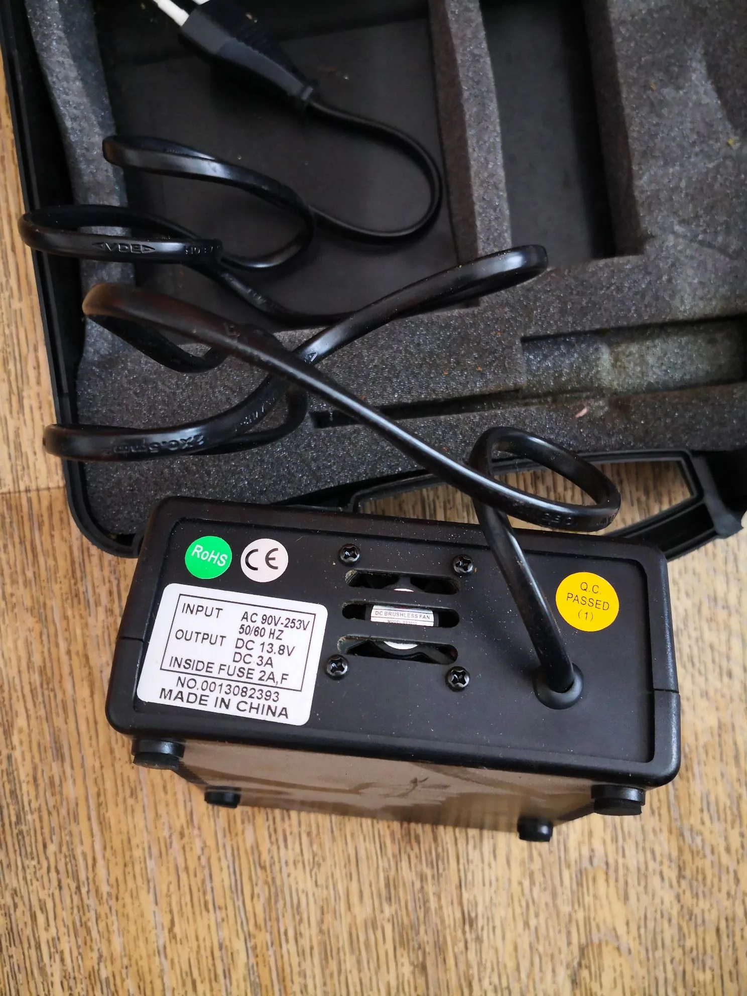 Czarny zasilacz sieciowy z naklejką techniczną, leżący w czarnym etui z piankową wkładką. Na białej etykiecie widnieje tekst: 'INPUT AC 90V-253V 50/60 HZ', 'OUTPUT DC 13.8V DC 3A', 'INSIDE FUSE 2A,F', 'NO.0013082393', 'MADE IN CHINA'. Obok znajdują się znaki certyfikacji RoHS i CE oraz żółna okrągła naklejka z napisem 'Q.C. PASSED'. Do zasilacza podłączony jest czarny kabel zasilający.