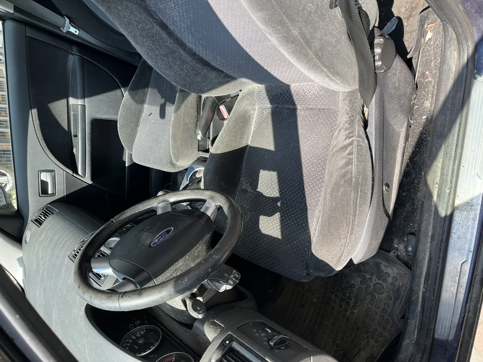 Wnętrze samochodu z widoczną kierownicą marki Ford, szarymi tkaninowymi fotelami, deską rozdzielczą z licznikami oraz otwartą drzwiami po stronie kierowcy. Na fotelach widoczne są cienie, a na podłodze leży czarna gumowa mata. Wnętrze pokryte jest kurzem i drobnymi zabrudzeniami. Drzwi posiadają uchwyt oraz przyciski sterujące szybą.