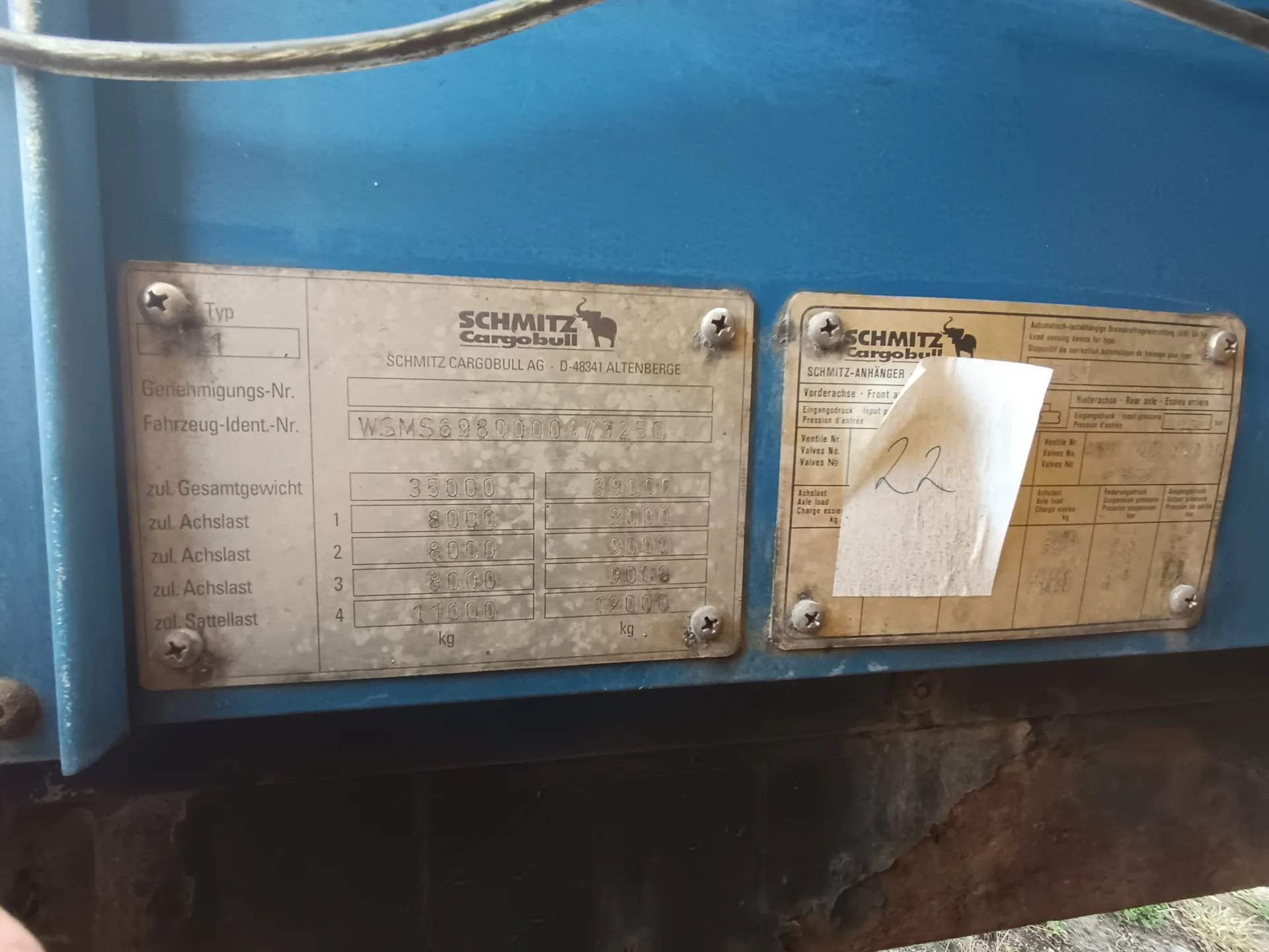 Dwa metalowe tablice identyfikacyjne na niebieskim tle. Lewa tablica z logo 'SCHMITZ Cargobull' i napisem 'SCHMITZ CARGOBULL AG - D-48341 ALTENBERGE', numerem identyfikacyjnym 'WSMS6980000473250', danymi o dopuszczalnych wagach: 'zul. Gesamtgewicht 35000 kg', 'zul. Achslast' dla osi 1-4 (8000, 8000, 8000, 11000 kg) oraz 'zul. Sattellast 12000 kg'. Prawa tablica z logo 'SCHMITZ Cargobull' i częściowo zakryta białą kartką z ręcznie zapisaną cyfrą '22'. W dolnej części obrazu widoczne są ślady rdzy na metalowej powierzchni.