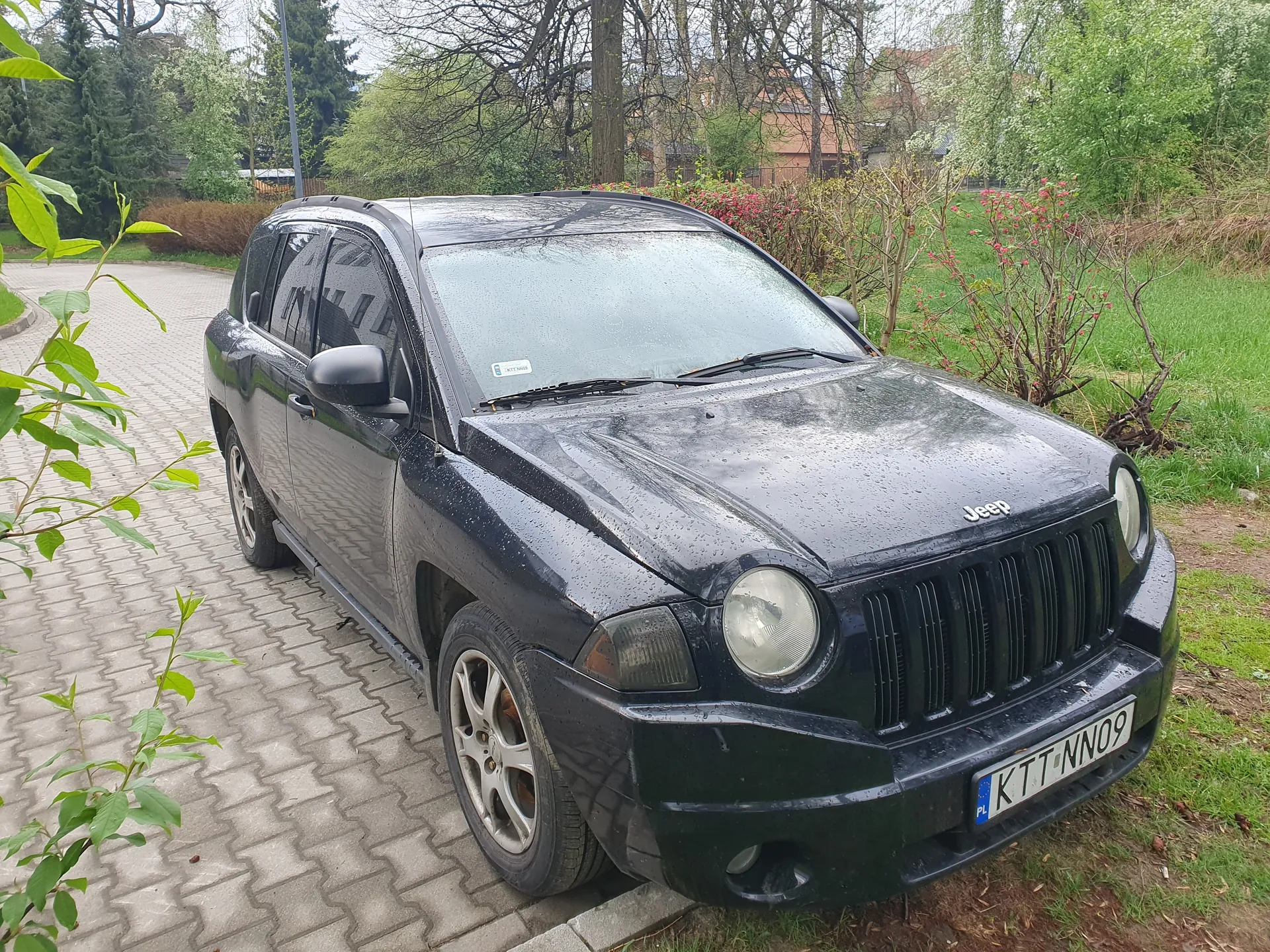 Czarny samochód marki Jeep z tablicą rejestracyjną KTT NN09 (PL) zaparkowany na brukowanej powierzchni. Na przedniej szybie widoczny jest mały naklejony znaczek. Samochód pokryty jest kroplami deszczu. W tle zielone krzewy, drzewa oraz budynki. Po lewej stronie obrazu rosną liście drzewa.