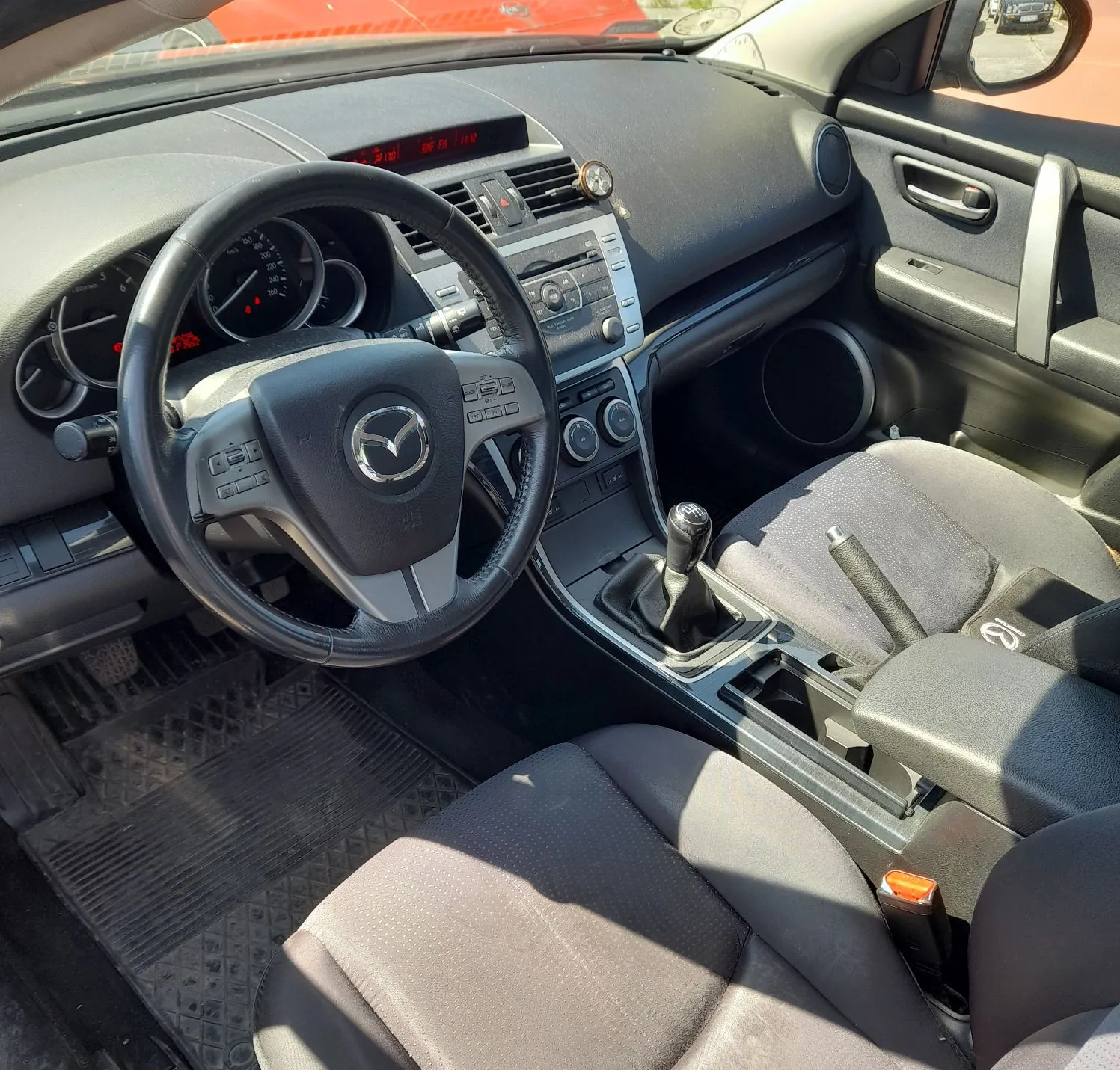 Wnętrze samochodu marki Mazda, widok z perspektywy kierowcy. Widoczna czarna kierownica z logo Mazdy w centrum, deska rozdzielcza z zegarami, systemem audio oraz klimatyzacją. Wnętrze wykończone w ciemnych kolorach, z szarymi materiałowymi fotelami. Na górze deski rozdzielczej znajduje się mały cyfrowy zegar z czerwonymi cyframi oraz dodatkowy analogowy wskaźnik. Widoczna lewarka zmiany biegów oraz podłogowa mata gumowa.