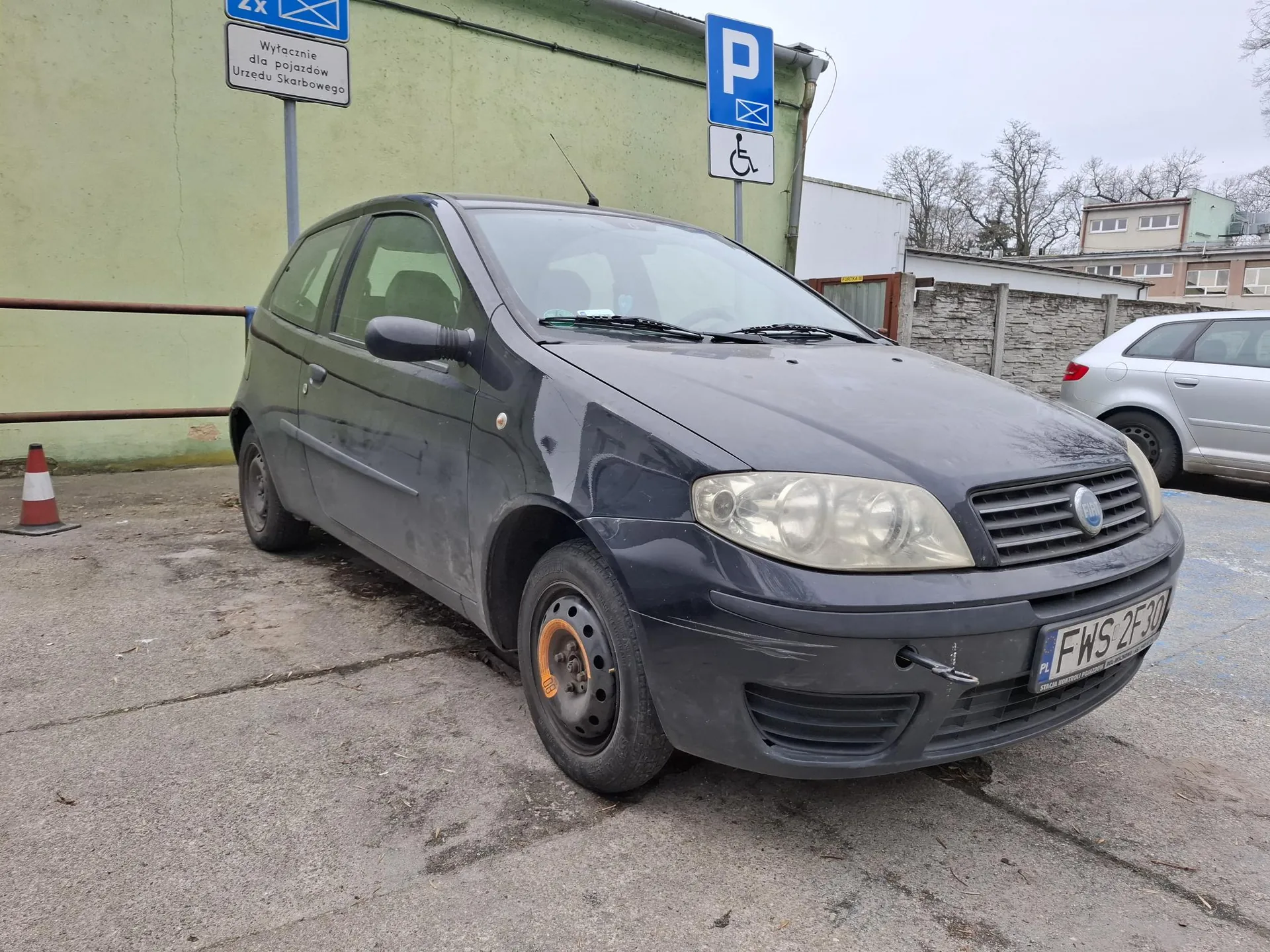 Ciemnoszary samochód Fiat zaparkowany na betonowym placu. Widoczna tablica rejestracyjna FWS 2F30. W tle zielona ściana budynku, niebieskie znaki parkingowe z literą P i symbolem niepełnosprawności, biały znak z napisem 'Wyłącznie dla pojazdów Urzędu Skarbowego' oraz pomarańczowo-biały stożek drogowy. Po prawej stronie częściowo widoczny srebrny samochód. Tło z drzewami i budynkami.