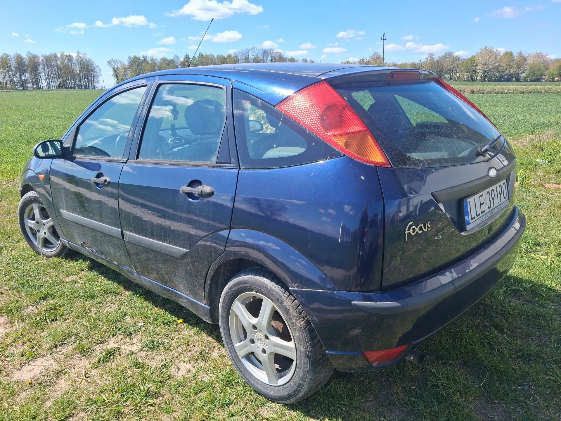 Granatowy samochód marki Ford Focus zaparkowany na trawiastym terenie. Pojazd jest widoczny z tyłu i z boku, posiada tablicę rejestracyjną o numerze LLE 39190. Nad samochodem widać błękitne niebo z białymi chmurami oraz linię drzew w oddali.