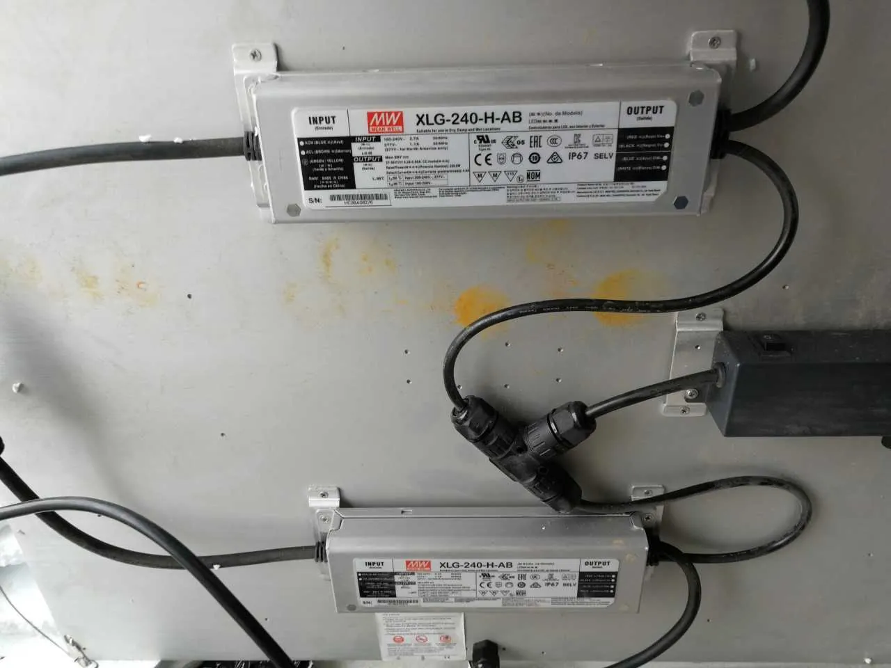 Na szarej metalowej powierzchni zamontowano dwa srebrne urządzenia elektroniczne oznaczone jako XLG-240-H-AB marki Mean Well (MW). Każdy z nich posiada etykietę z sekcjami INPUT i OUTPUT, symbolami certyfikacji (m.in. CE, IP67, SELV) oraz czarnymi kablami podłączonymi do złączy. Na powierzchni widoczne są żółtawe plamy i śruby mocujące urządzenia.