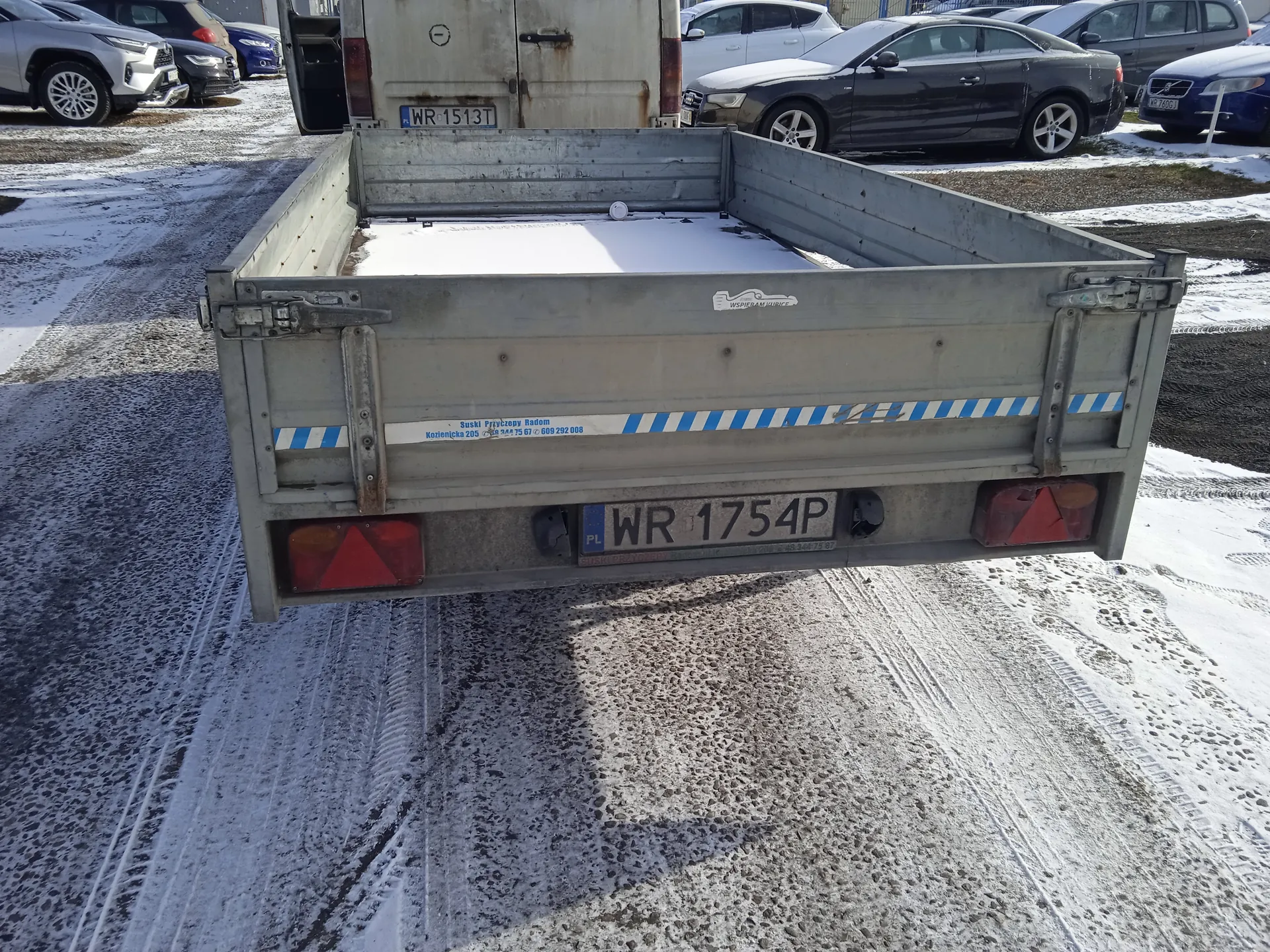Tył ciężarówki z pustą, częściowo zasłaną śniegiem naczepą. Widoczny polski numer rejestracyjny WR 1754P oraz naklejka z napisem „Słupsk Przyczepy Radom, Koszalińska 205, tel. 504 75 67, 609 292 008”. W tle zaparkowane samochody, w tym pojazd z numerem WR 1513T, oraz śnieżna droga z odciskami opon.