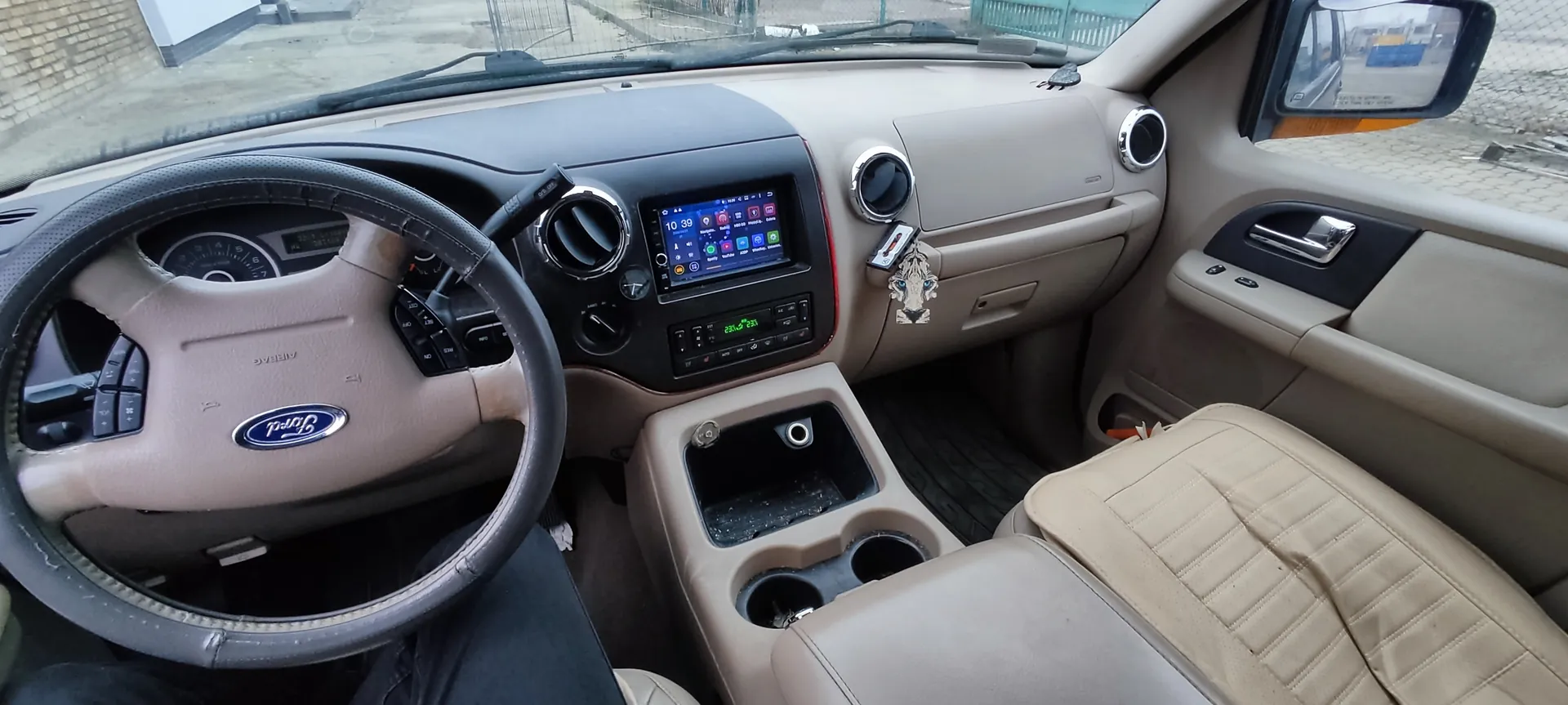 Wnętrze samochodu marki Ford z beżową tapicerką. Widoczna kierownica z logo Ford, deska rozdzielcza z cyfrowym wyświetlaczem pokazującym godzinę 10:39 oraz ikony aplikacji. W centralnej konsoli znajduje się ekran multimedialny, przyciski sterujące i uchwyty na napoje. Na desce rozdzielczej wiszą kluczyki z ozdobą w kształcie głowy lwa. Przez przednią szybę widać fragment budynku i ogrodzenie. Siedzenia są wykończone beżową skórą z przeszyciami.