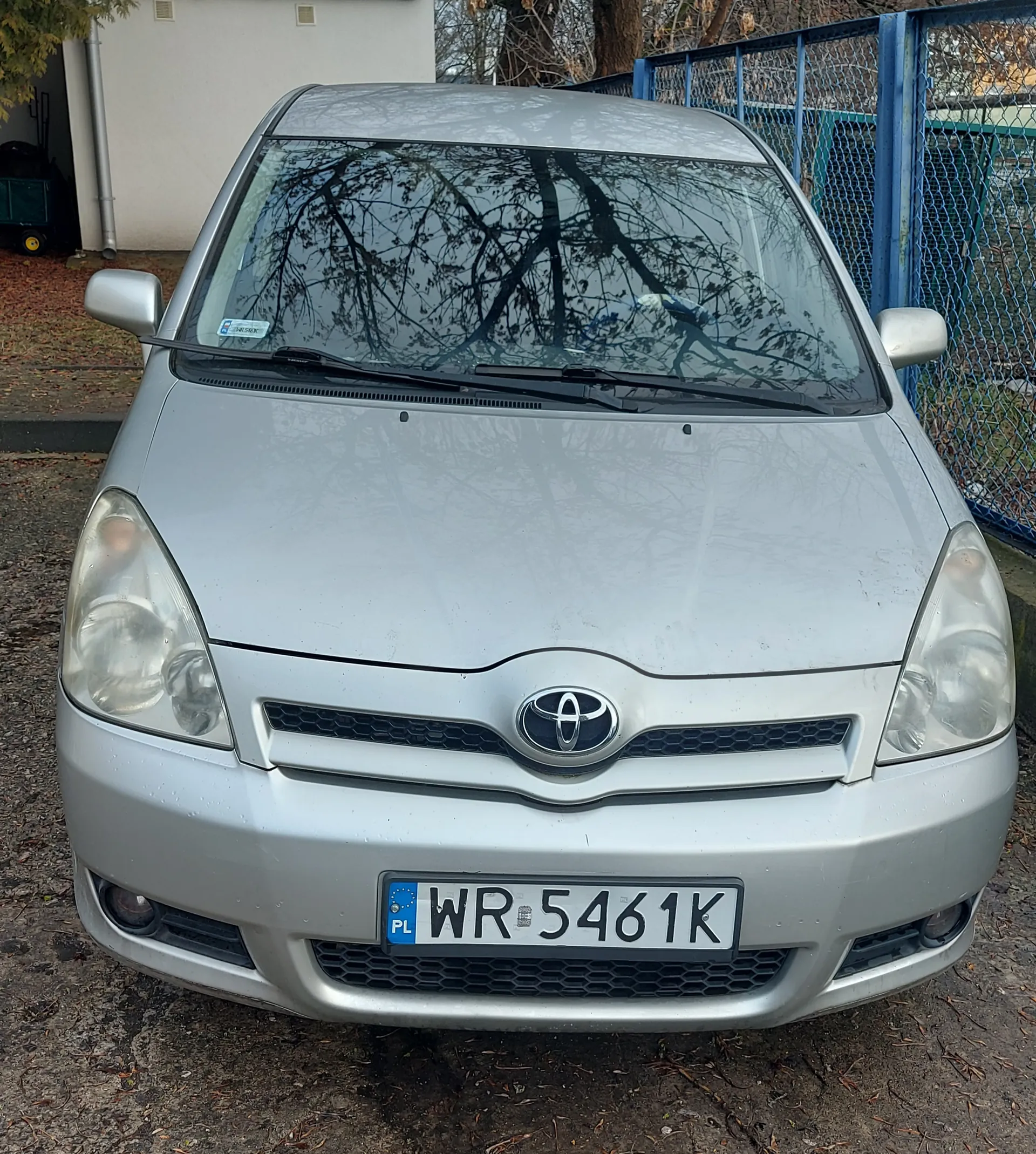 Przód srebrnego samochodu marki Toyota z tablicą rejestracyjną PL WR 5461K. Tło: niebieskie ogrodzenie, budynek z białą ścianą, drzewa bez liści i ziemia z suchymi liśćmi. Odbicie drzew w przedniej szybie.