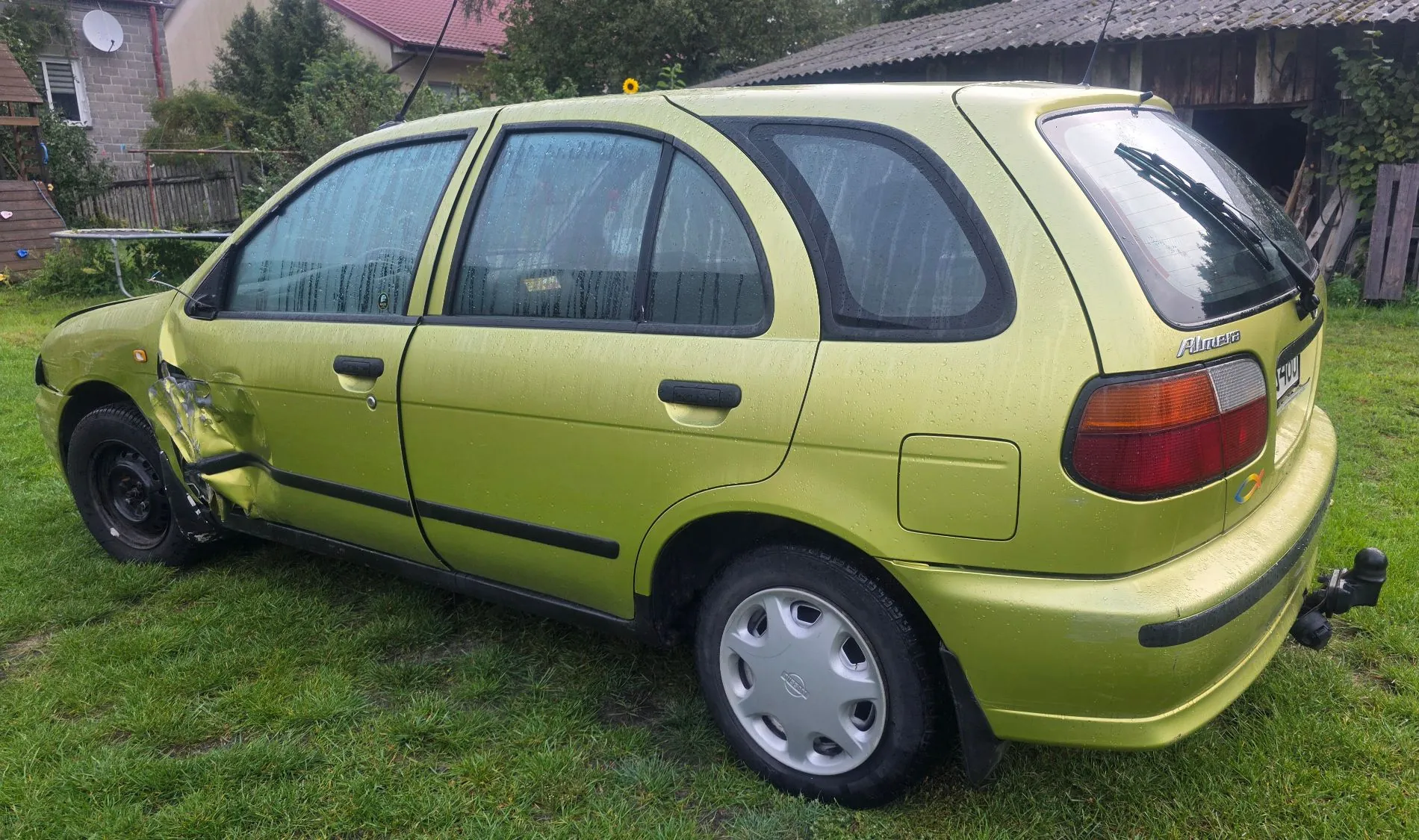 Jasnozielony samochód marki Nissan Almera w nadwoziu hatchback, zaparkowany na trawie. Pojazd posiada widoczne uszkodzenia karoserii w postaci wgniecenia na przednim lewym błotniku i drzwiach kierowcy. Na tylnej klapie widoczny jest napis 'Almera'. Samochód jest wyposażony w hak holowniczy.
