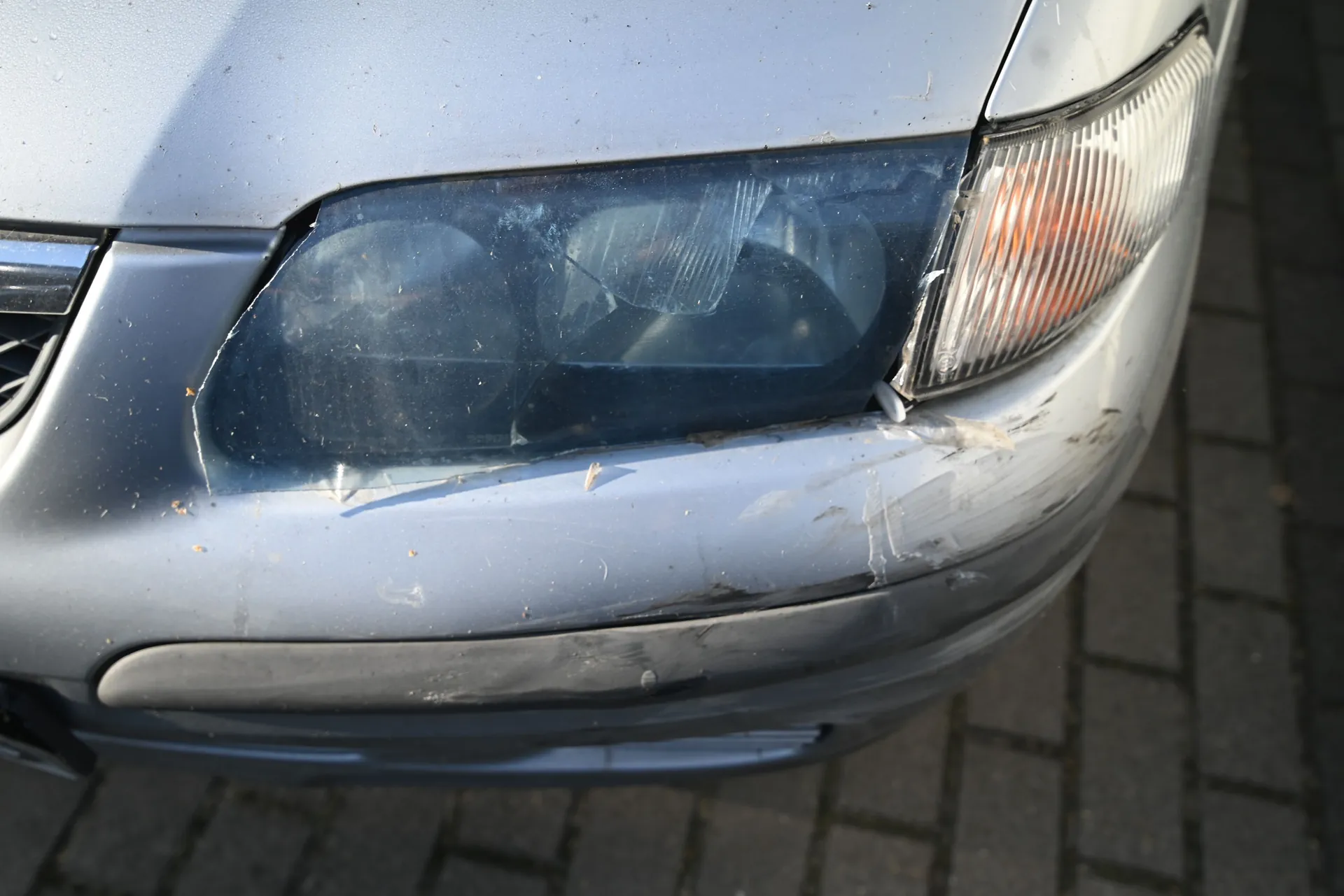 Zbliżenie na przedni lewy reflektor i zderzak srebrnego samochodu. Reflektor jest pęknięty i zmatowiały, a na lakierze zderzaka poniżej lampy widoczne są liczne zarysowania i odpryski. Samochód stoi na kostce brukowej.