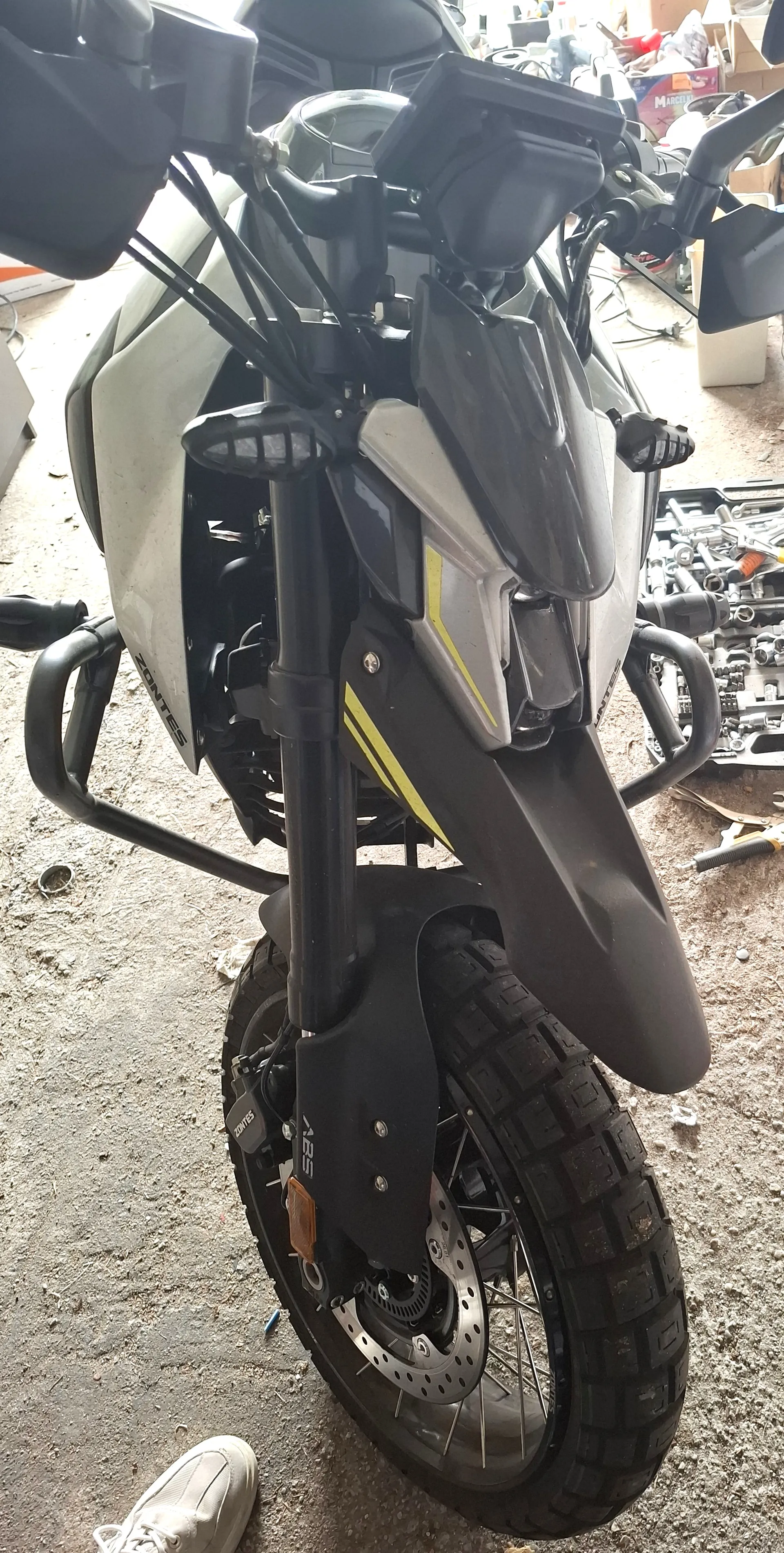 Przednia część motocykla marki ZONTES w kolorze białym z czarnymi i żółtymi akcentami. Widoczne są czarne uchwyty, lusterka, przednie koło z grubą oponą, hamulec tarczowy oraz napisy 'ZONTES' na boku i 'ABS' na czarnej osłonie. W tle warsztat z narzędziami, częściami mechanicznymi i białym butem na podłodze. Podłoga jest brudna, z widocznymi śladami kurzu i śmieci.