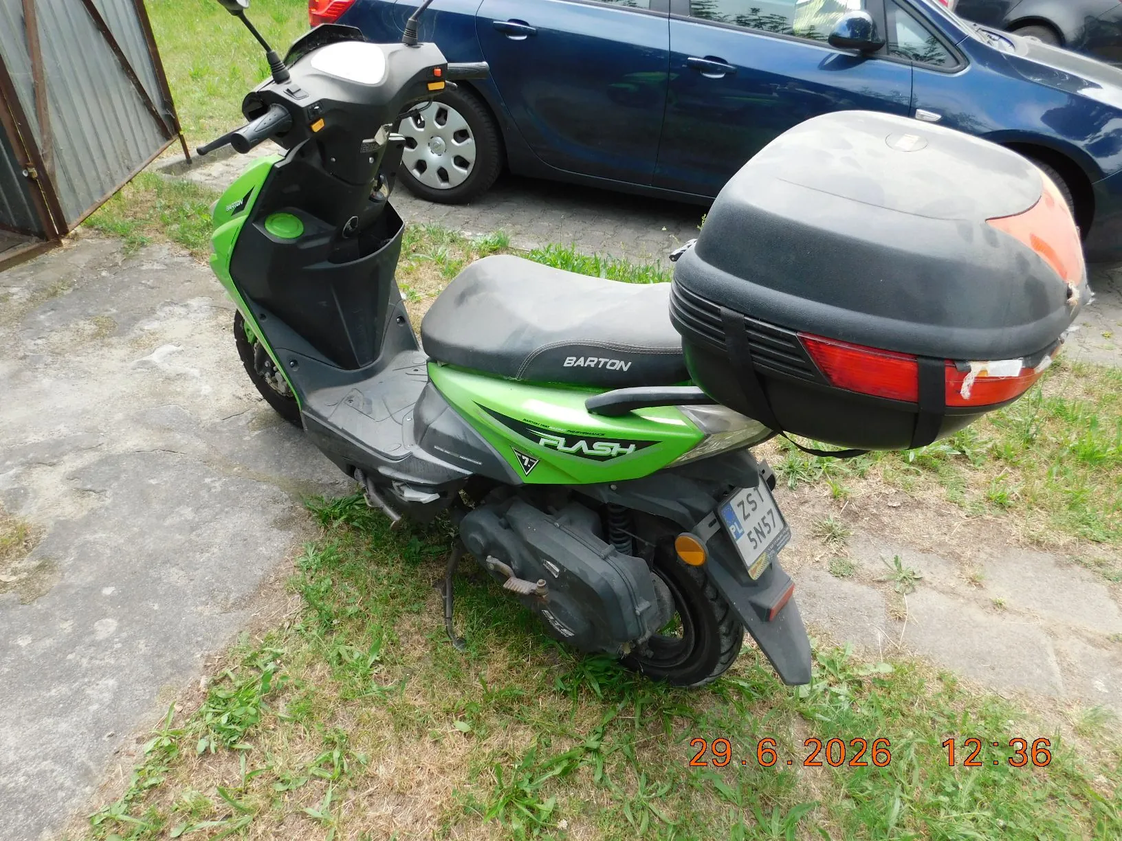 Zielony skuter marki Barton Flash z zamontowanym czarnym kufrem z tyłu. Pojazd stoi na betonowym podjeździe z kępami trawy. W tle widoczny jest fragment niebieskiego samochodu osobowego oraz metalowa konstrukcja. Na tylnej tablicy rejestracyjnej widnieje napis ZS 5N57. W prawym dolnym rogu zdjęcia znajduje się czerwony datownik z tekstem 29.6.2026 12:36.