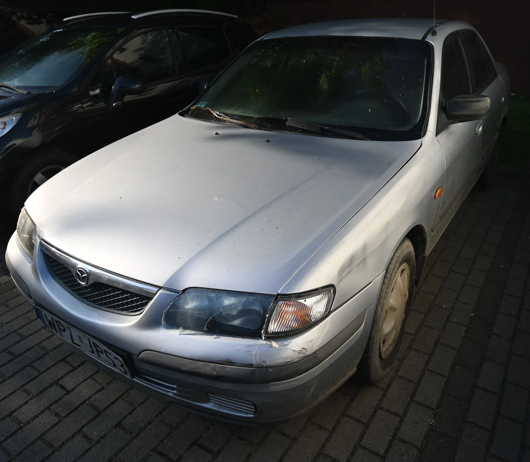 Srebrny samochód osobowy marki Mazda zaparkowany na kostce brukowej. Pojazd ma widoczne uszkodzenia na przednim zderzaku oraz masce. W tle widoczny jest fragment innego, czarnego samochodu. Tablica rejestracyjna zaczyna się od liter WPL.