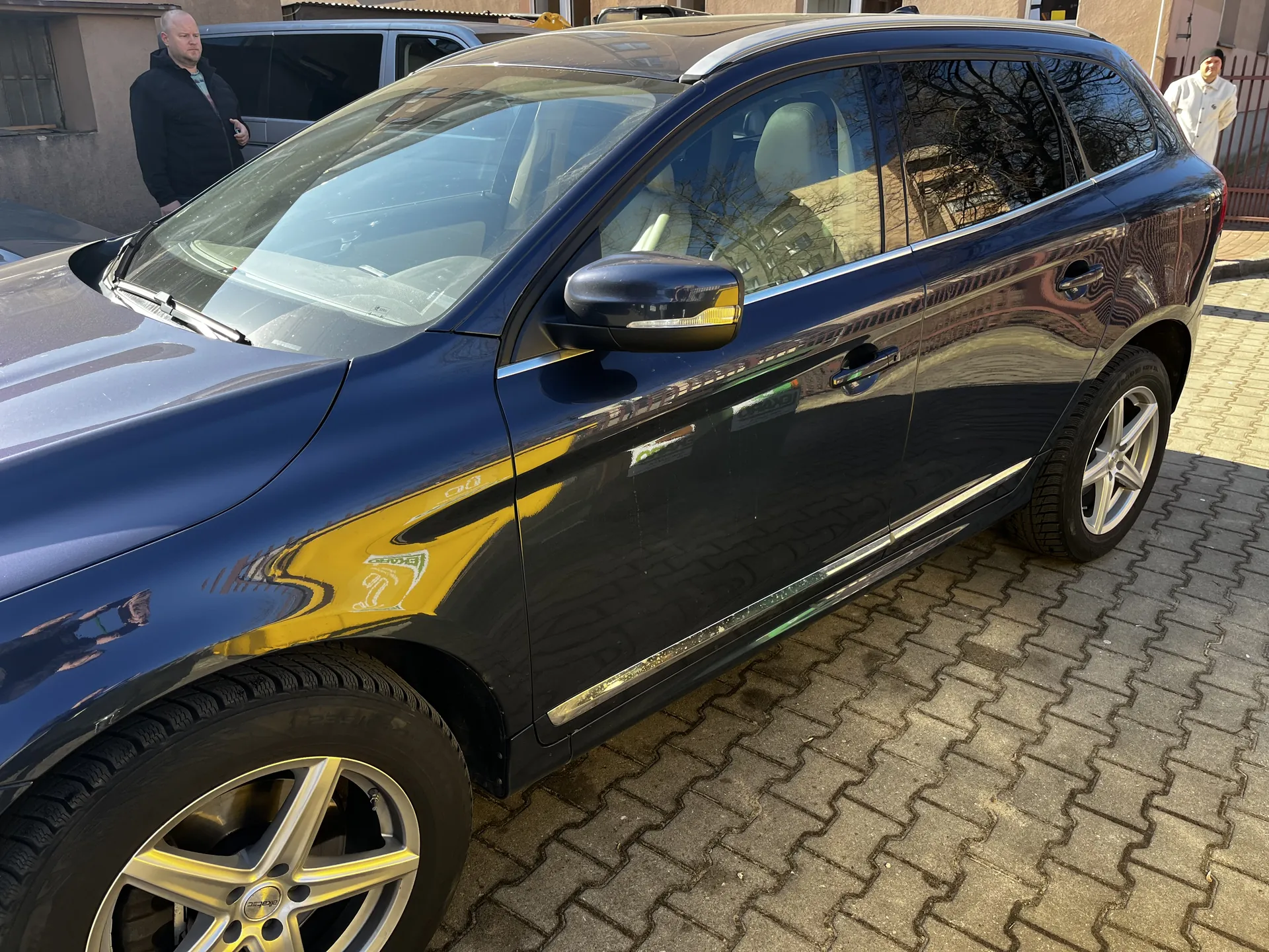 Ciemnoniebieski SUV zaparkowany na brukowanej powierzchni. Widoczne są przednie i boczne części pojazdu, w tym felgi ze srebrnymi dyskami. W tle stoją dwie osoby: jedna w czarnej kurtce blisko samochodu, druga w białej kurtce w oddali. Za nimi budynki z oknami i ogrodzenie. Odbicia na karoserii wskazują na słoneczny dzień.