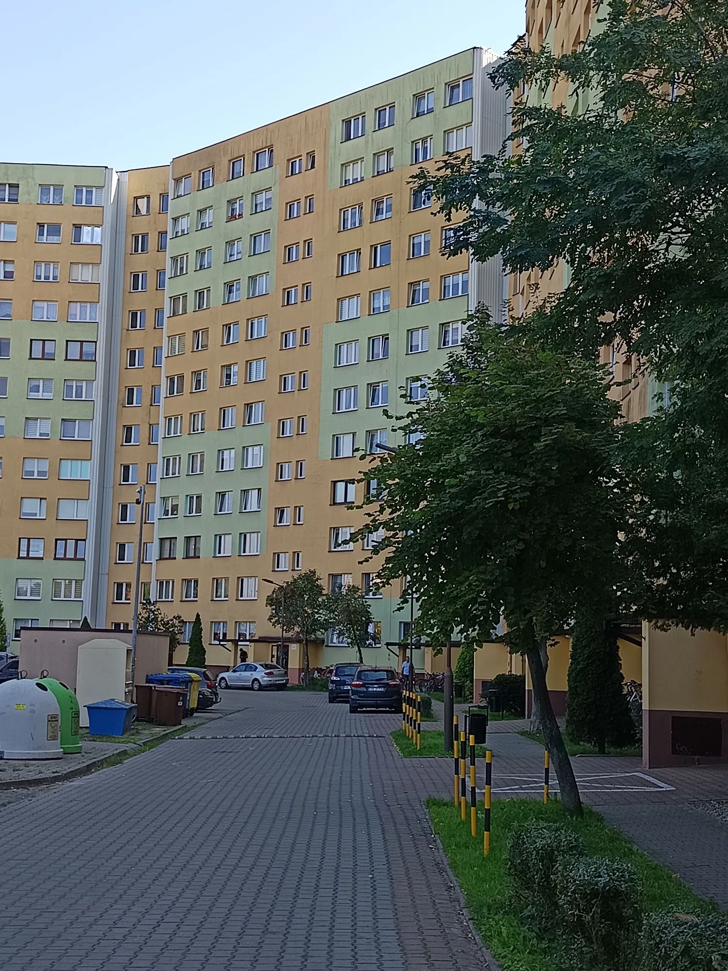 Wielopiętrowy blok mieszkalny z elewacją w kolorach żółtym i jasnozielonym. Przed budynkiem znajduje się brukowana droga dojazdowa, zaparkowane samochody oraz zieleń w postaci drzew i krzewów. Po lewej stronie widoczne są kontenery na odpady.