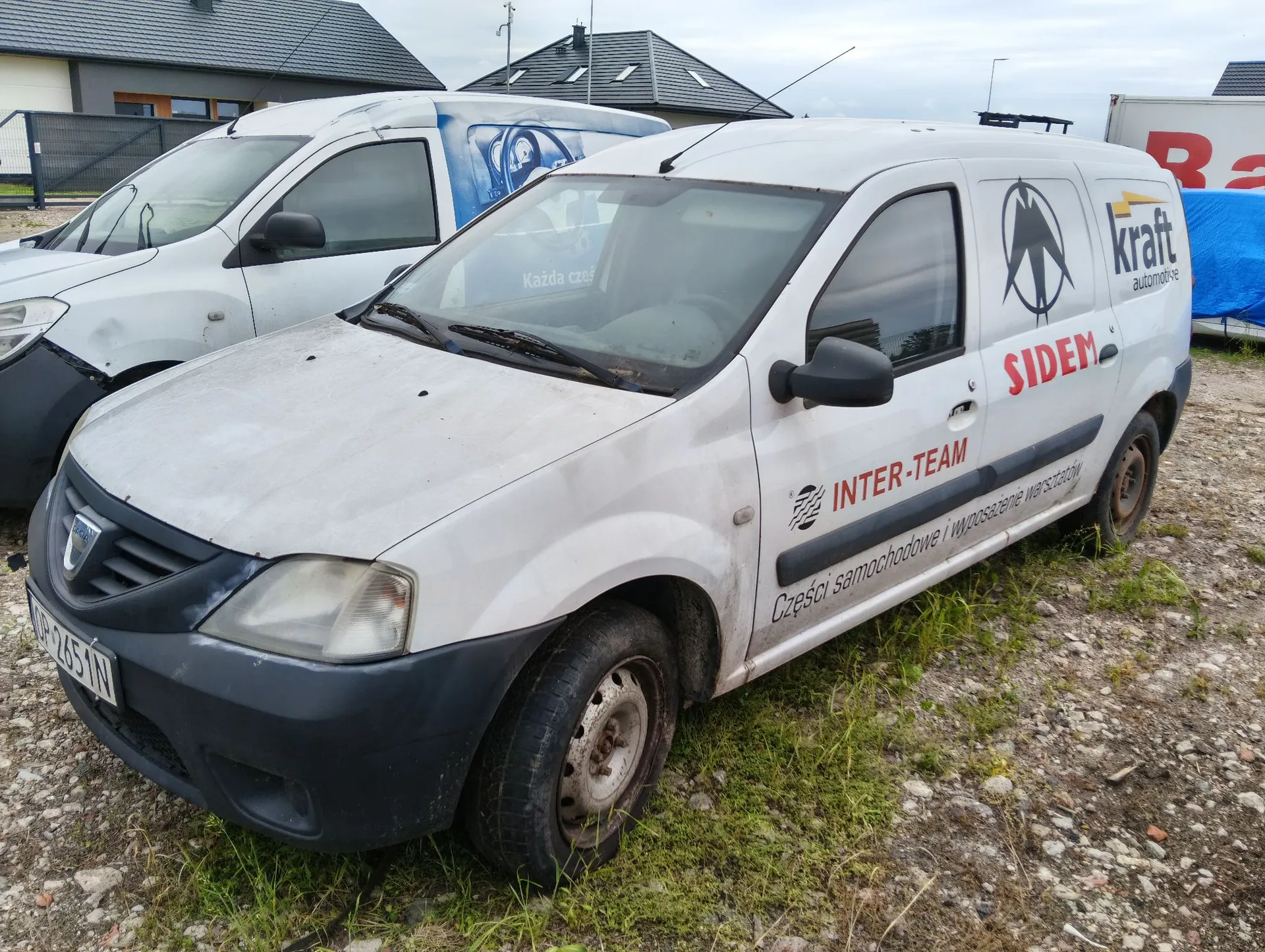 Biały samochód dostawczy marki Dacia zaparkowany na żwirowym podłożu. Na boku pojazdu znajdują się napisy: 'INTER-TEAM', 'Części samochodowe i wyposażenie warsztatów' oraz logo 'kraft automotive' i napis 'SIDEM'. W tle widoczny jest drugi, biały samochód dostawczy oraz fragmenty budynków z ciemnymi dachami.