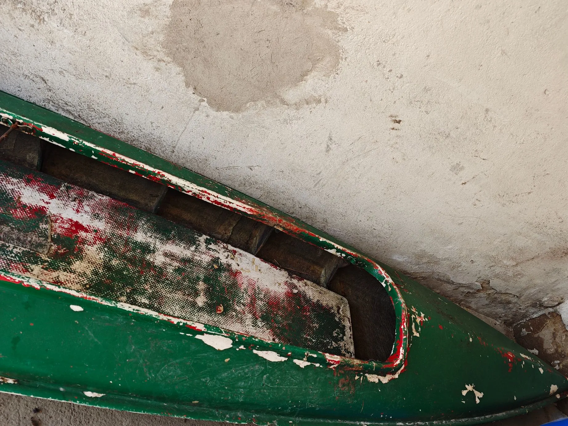 Zbliżenie na fragment starej, zielonej łodzi z widocznymi uszkodzeniami lakieru i czerwonymi przetarciami wewnątrz kadłuba. Łódź opiera się o jasną, chropowatą ścianę o tynkowanej strukturze.