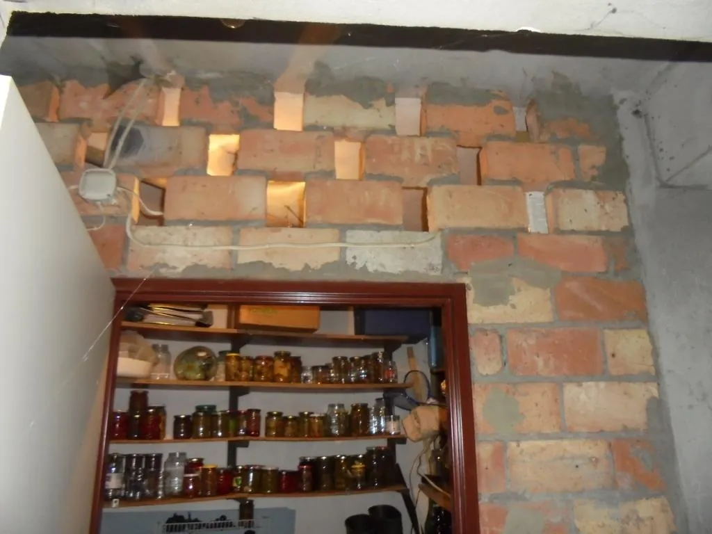 Ceglana ściana z brakującymi cegiełkami tworzącymi prostokątne otwory, nad drewnianą półką z różnymi słoikami i pojemnikami. Na ścianie widoczne są białe przewody i mały biały pudełko elektryczne. Półka z brązowym ramieniem zawiera wiele rzędów słoików z różnymi zawartościami, a na górnej półce znajdują się pudełka i inne przedmioty. Fragment białej ściany po lewej stronie i sufitu na górze.