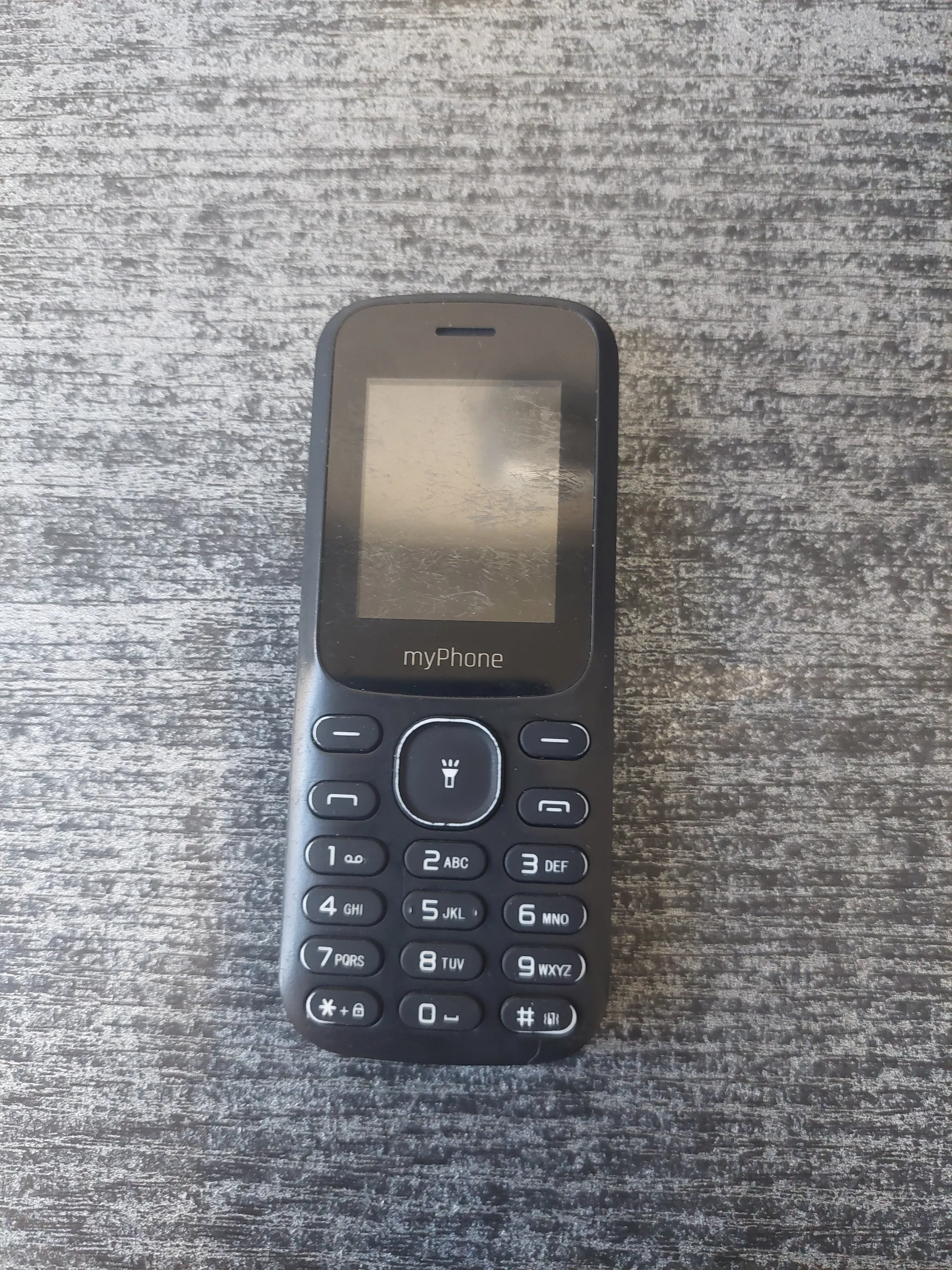Czarny telefon komórkowy marki myphone z ekranem i klawiaturą numeryczną. Klawisze z cyframi od 1 do 9, 0, * i # oraz literami (np. 2 ABC, 3 DEF, 4 GHI). Telefon leży na szarym, teksturowanym tle.