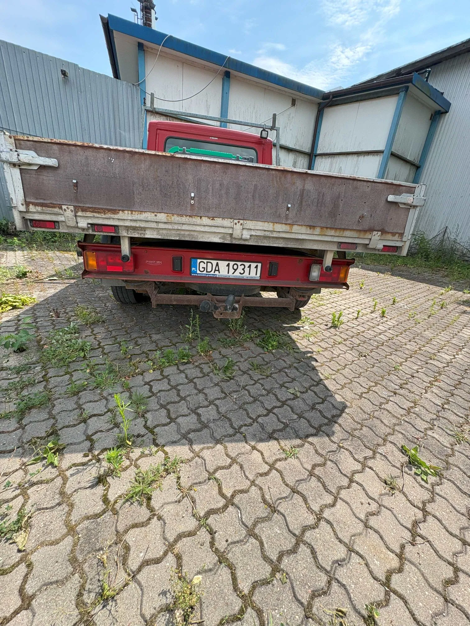 Tylny widok czerwonego samochodu ciężarowego z otwartą skrzynią ładunkową, zaparkowanego na kostce brukowej. Pojazd posiada polską tablicę rejestracyjną o numerze GDA 19311. W tle widoczny jest budynek przemysłowy z niebieskimi elementami konstrukcyjnymi i jasnymi ścianami.