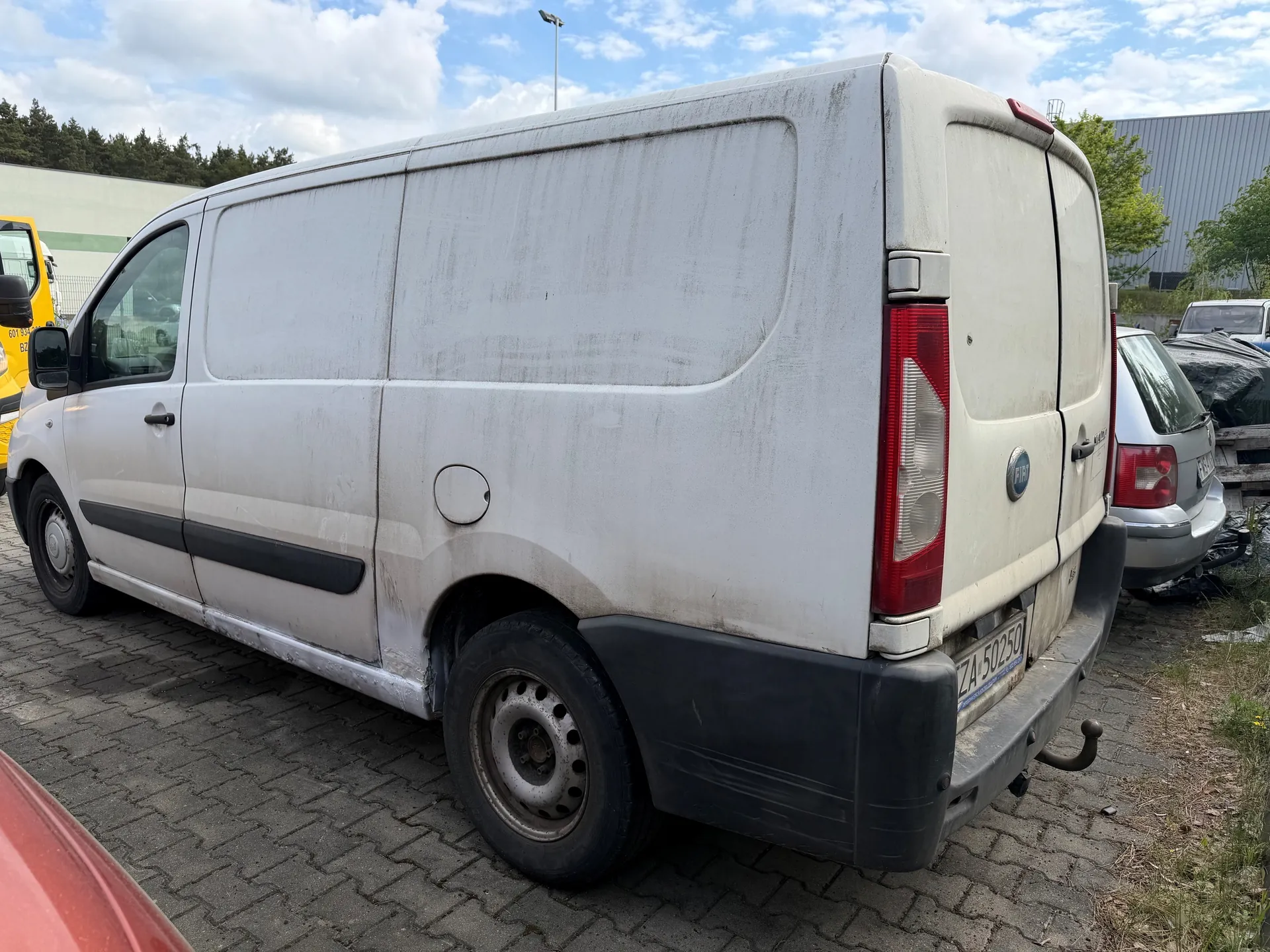 Biały dostawczy samochód Fiat z widoczną tablicą rejestracyjną ZA 50250, zaparkowany na brukowanym placu. Pojazd ma zabrudzoną karoserię, czarną listwę boczną i czarny zderzak tylny. W tle widoczne są inne pojazdy: żółty samochód po lewej stronie, srebrny samochód osobowy po prawej oraz budynki przemysłowe. Na niebie znajdują się chmury, a w oddali rosną drzewa.