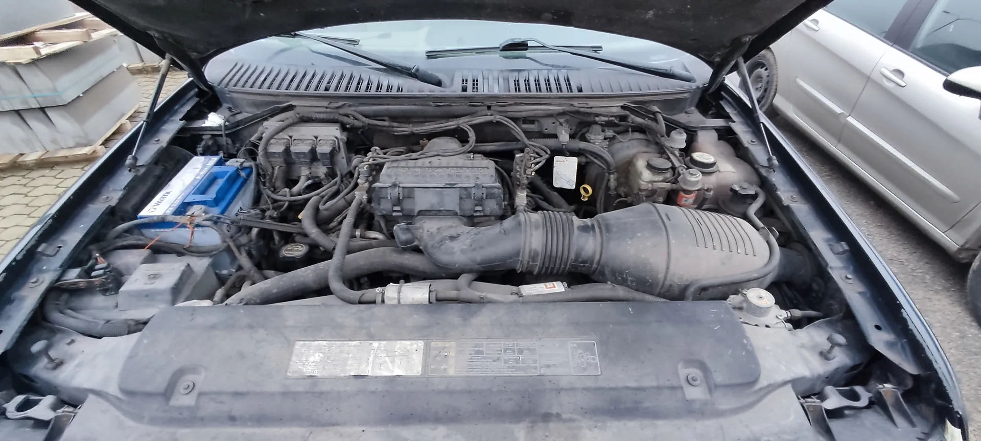 Zdjęcie przedstawia otwarty kapotę samochodu z widocznym silnikiem. W komorze silnika znajdują się różne elementy: niebieski akumulator z napisem VARTA, czarne przewody, rury, zbiorniki oraz plastikowa pokrywa z naklejkami. W tle widoczny jest srebrny samochód zaparkowany obok oraz stosy betonowych elementów na paletach. Podłoga wyłożona jest kostką brukową.