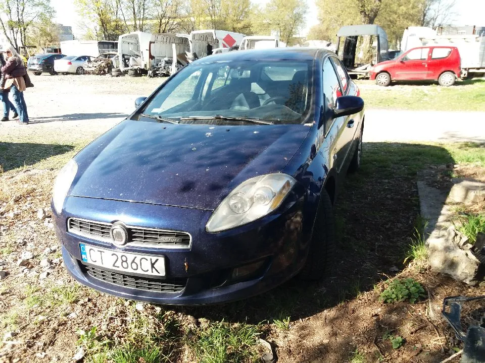 Ciemnoniebieski samochód osobowy z tablicą rejestracyjną CT 286CK zaparkowany na terenie z trawą i kamieniami. W tle widoczne są inne pojazdy, w tym czerwony samochód i przyczepy, oraz dwie osoby idące po lewej stronie. Tło stanowią drzewa i budynki.