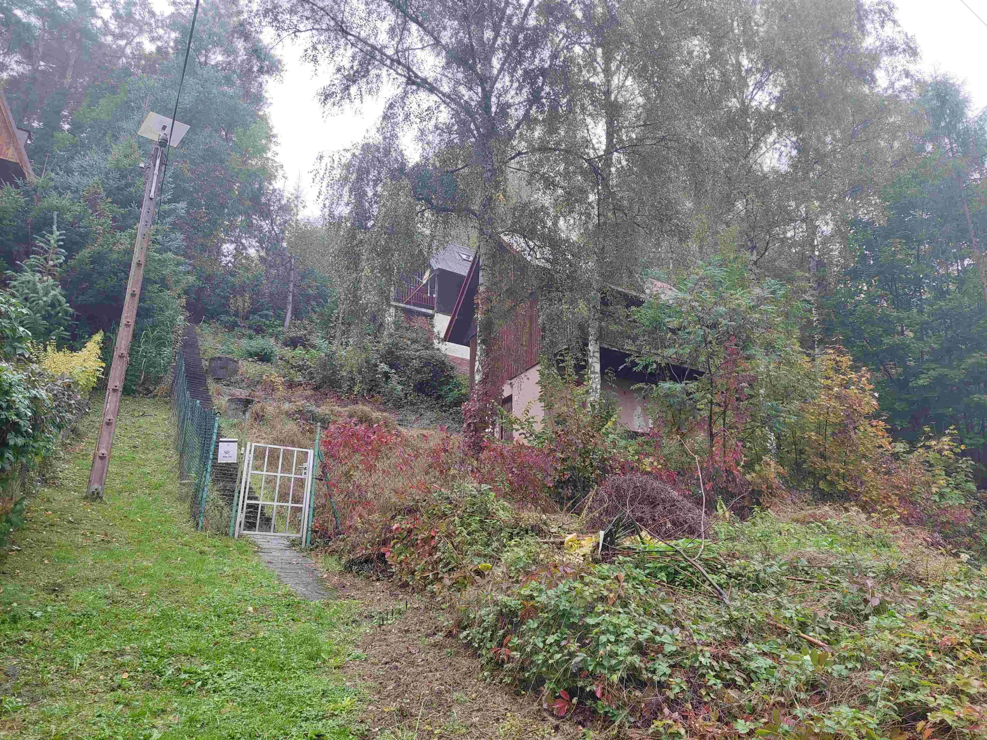 Dom na stromym zboczu otoczony gęstym lasem i roślinnością. W pierwszym planie trawiasty teren z metalową bramą oraz zielonym ogrodzeniem. Po lewej stronie widoczny drewniany słup energetyczny z panelem słonecznym. Dom ma pochylony dach i jest częściowo zasłonięty drzewami. Na bramie biała tabliczka z nieczytelnym napisem. Wokół rosną krzewy z zielonymi i jesiennymi liśćmi, a na ziemi leżą suche gałęzie. Niebo jest zachmurzone.