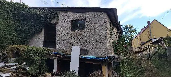 Stara, dwupiętrowa kamienica z teksturowaną elewacją, częściowo porośnięta pnączami. Na pierwszym piętrze widoczne są dwa otwory: okno i puste przejście. Na parterze znajduje się przedsionek z niebieską folią i białym pionowym elementem. W tle żółty dom z czerwonym dachem, czarny metalowy płot oraz zarośla. Niebo częściowo zachmurzone. Wokół budynku rozrzucone są materiały budowlane i porosty.