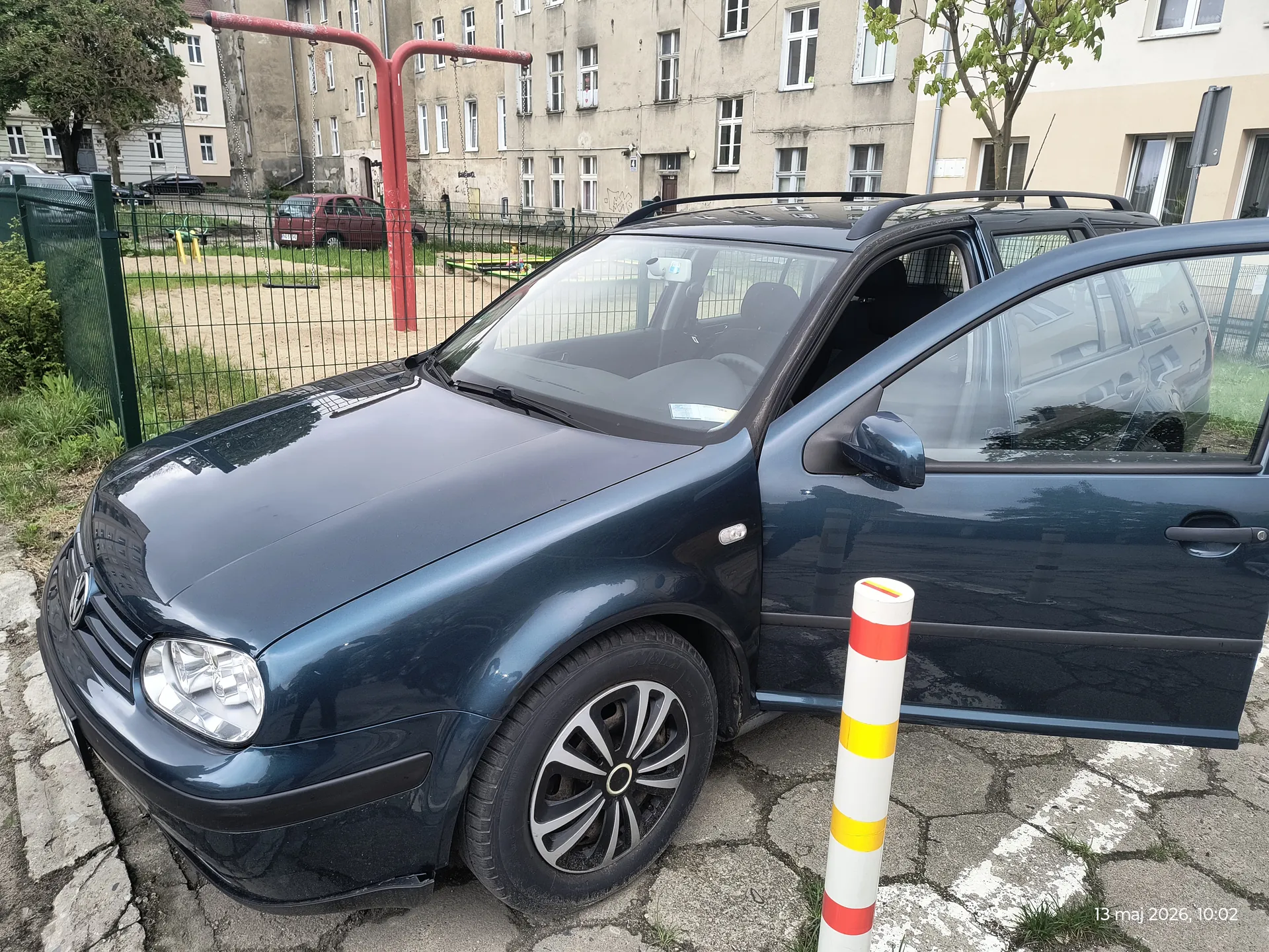 Ciemnoniebieski samochód osobowy z otwartą przodem drzwiami zaparkowany na brukowanej powierzchni. Przy samochodzie stoi biało-czerwono-żółty słup. W tle zielone ogrodzenie, boisko z czerwonymi huśtawkami, budynki mieszkalne z oknami oraz czerwony samochód. W prawym dolnym rogu widoczny napis: 13 maj 2026, 10:02.