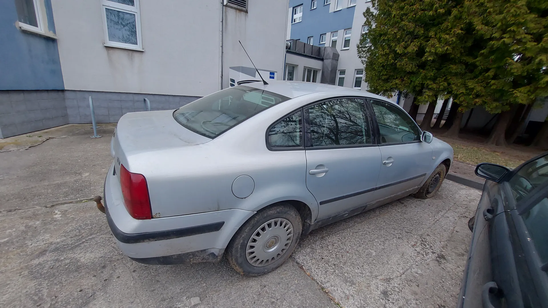 Srebrny samochód osobowy typu sedan zaparkowany na betonowej powierzchni. Pojazd ma zabrudzone koła i dolne części nadwozia. W tle widoczne są budynki z białymi i niebieskimi elewacjami, oknami oraz drzewo z zielonymi liśćmi. Po prawej stronie częściowo widoczny jest czarny samochód. Na betonie znajdują się pęknięcia i plamy.