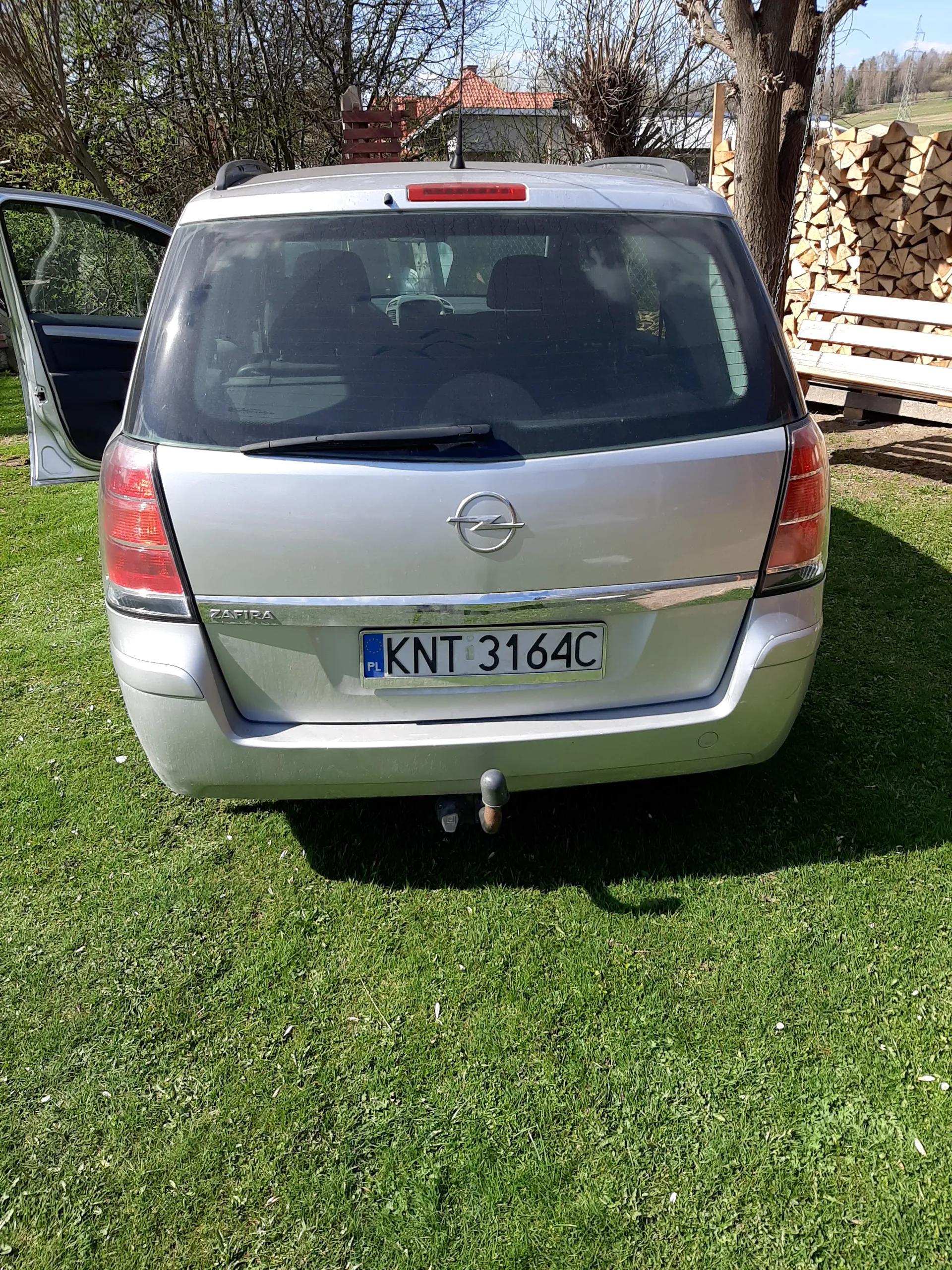 Srebrny samochód marki Opel Zafira z widocznym logo na tylnej klapie. Tablica rejestracyjna KNT 3164C z oznaczeniem PL. Drzwi kierowcy są otwarte. Samochód zaparkowany na zielonej trawie. W tle stos drewna, drzewa oraz dom z czerwonym dachem. Na tablicy rejestracyjnej widoczny jest niebieski pas z literą PL.