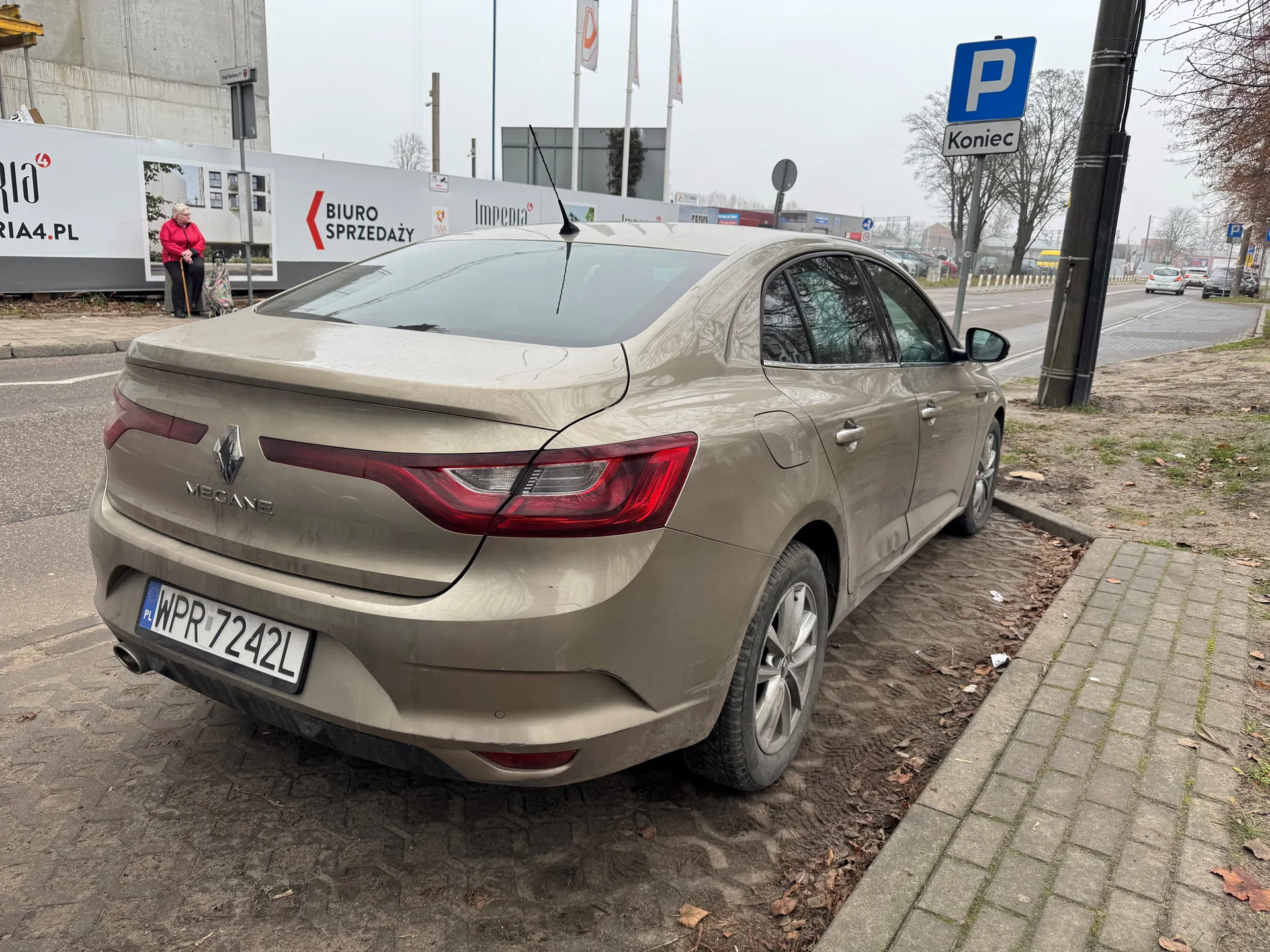 Zdjęcie przedstawia beżowy samochód marki Renault Megane zaparkowany na poboczu. Widoczny jest tył pojazdu z tablicą rejestracyjną NPR 7242L. W tle znajduje się niebieski znak parkingowy z literą P i napisem Koniec, budynek z transparentem BIURO SPRZEDAŻY oraz osoba w czerwonej kurtce stojąca przy barierce. Na drodze w oddali widoczne są inne pojazdy, a po prawej stronie chodnik z bruku.