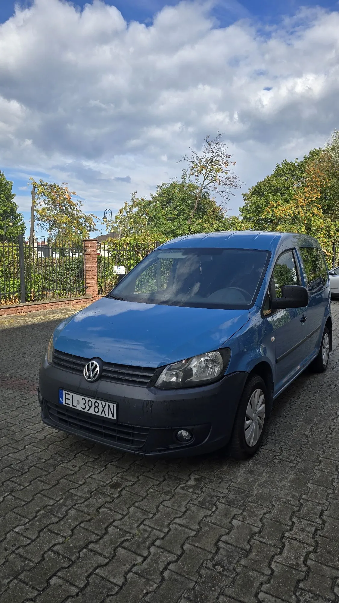 Niebieski samochód marki Volkswagen zaparkowany na kostce brukowej. Pojazd posiada polską tablicę rejestracyjną o numerze EL 398XN. W tle widoczny jest czarny metalowy płot, ceglany słupek oraz zielone drzewa pod zachmurzonym niebem.
