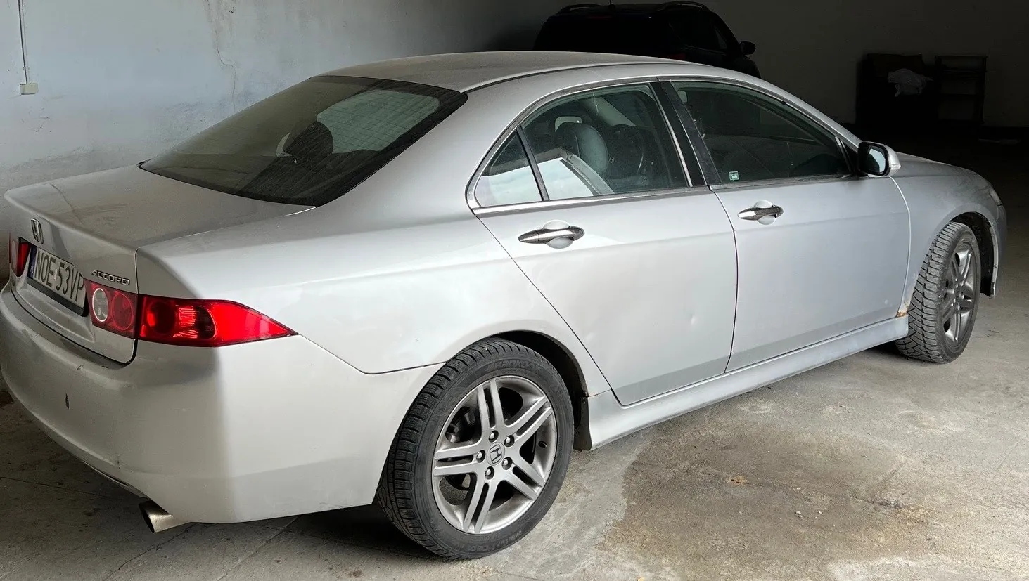 Srebrny samochód osobowy Honda Accord zaparkowany w garażu. Widoczna tablica rejestracyjna z numerem NOE 53VP, czerwone światła tylne, srebrne felgi oraz fragmenty betonowych ścian i podłogi. W tle ciemny samochód.