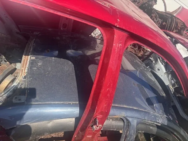 Zdjęcie przedstawia uszkodzony fragment samochodu w stanie wraku. Widoczna jest czerwona metalowa konstrukcja, prawdopodobnie część drzwi lub nadwozia, z widocznymi śladami uszkodzeń i zadrapań. W tle znajduje się niebieska część nadwozia z otwartym przestrzenią wewnętrzną, zawierającą elementy mechaniczne i zardzewiałe części. Na powierzchniach widoczne są ślady brudu i korozji. W tle rozpoznaje się fragmenty innych zniszczonych pojazdów oraz suchą trawę.