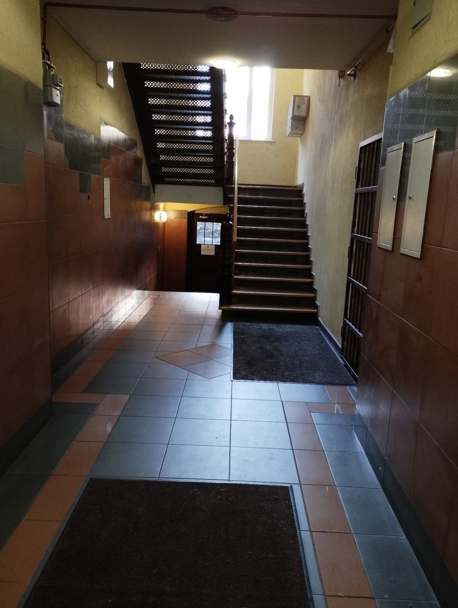 Wnętrze korytarza w budynku z kafelkami na ścianach i podłodze. Po prawej stronie znajdują się brązowe płytki ścienne, a po lewej ciemniejsze, szaro-brązowe. W głębi korytarza widoczne są drewniane schody prowadzące na wyższe piętro oraz ciemne drzwi. Na podłodze leżą dwie ciemne wycieraczki. Nad schodami znajduje się okno wpuszczające światło dzienne.